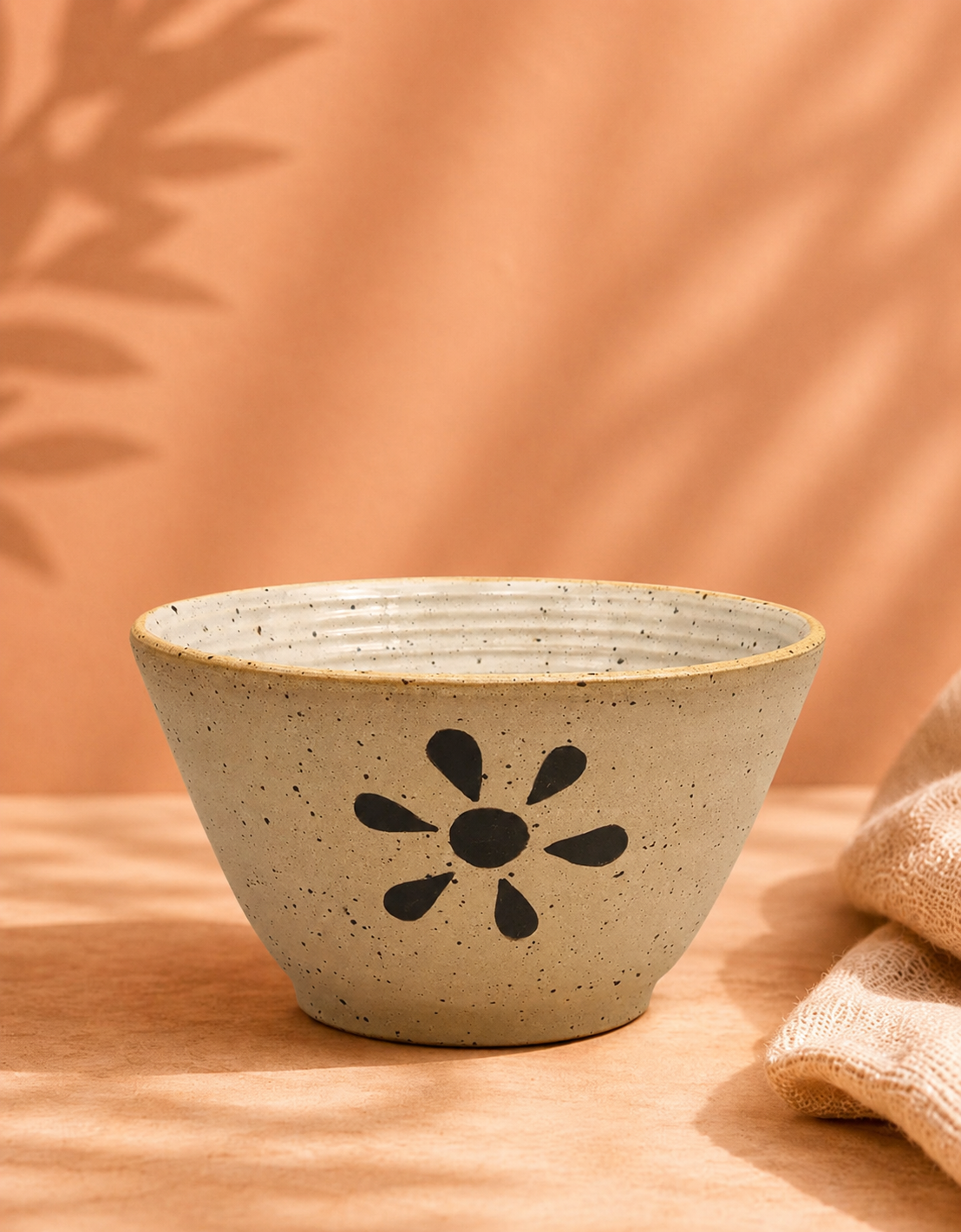 A ceramic bowl with a black flower design on a wooden surface, with a beige fabric and pink background.