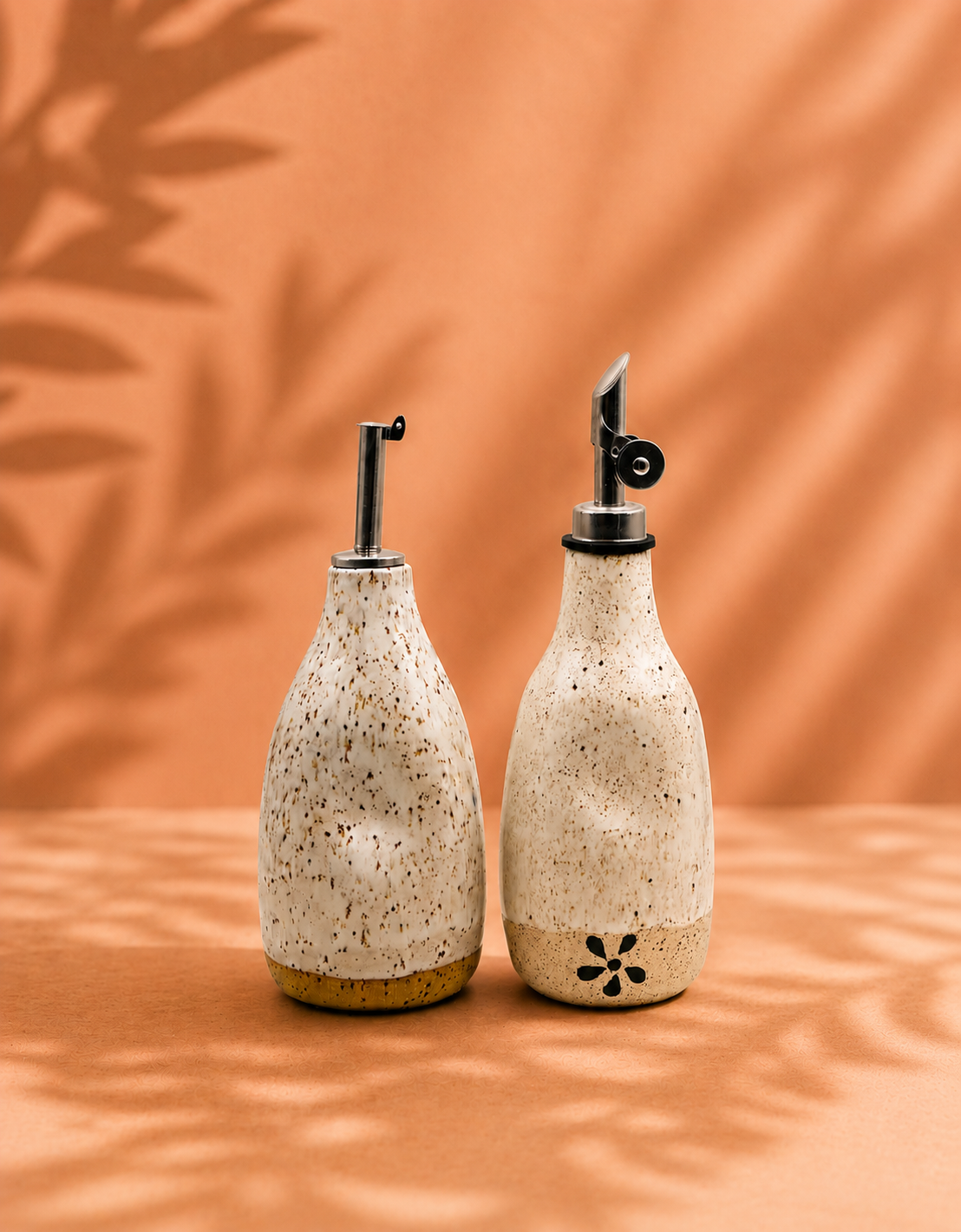Two speckled ceramic oil and vinegar dispensers with stainless steel pour spouts on a peach-colored surface, against a matching peach background with blurred leaf shadow.