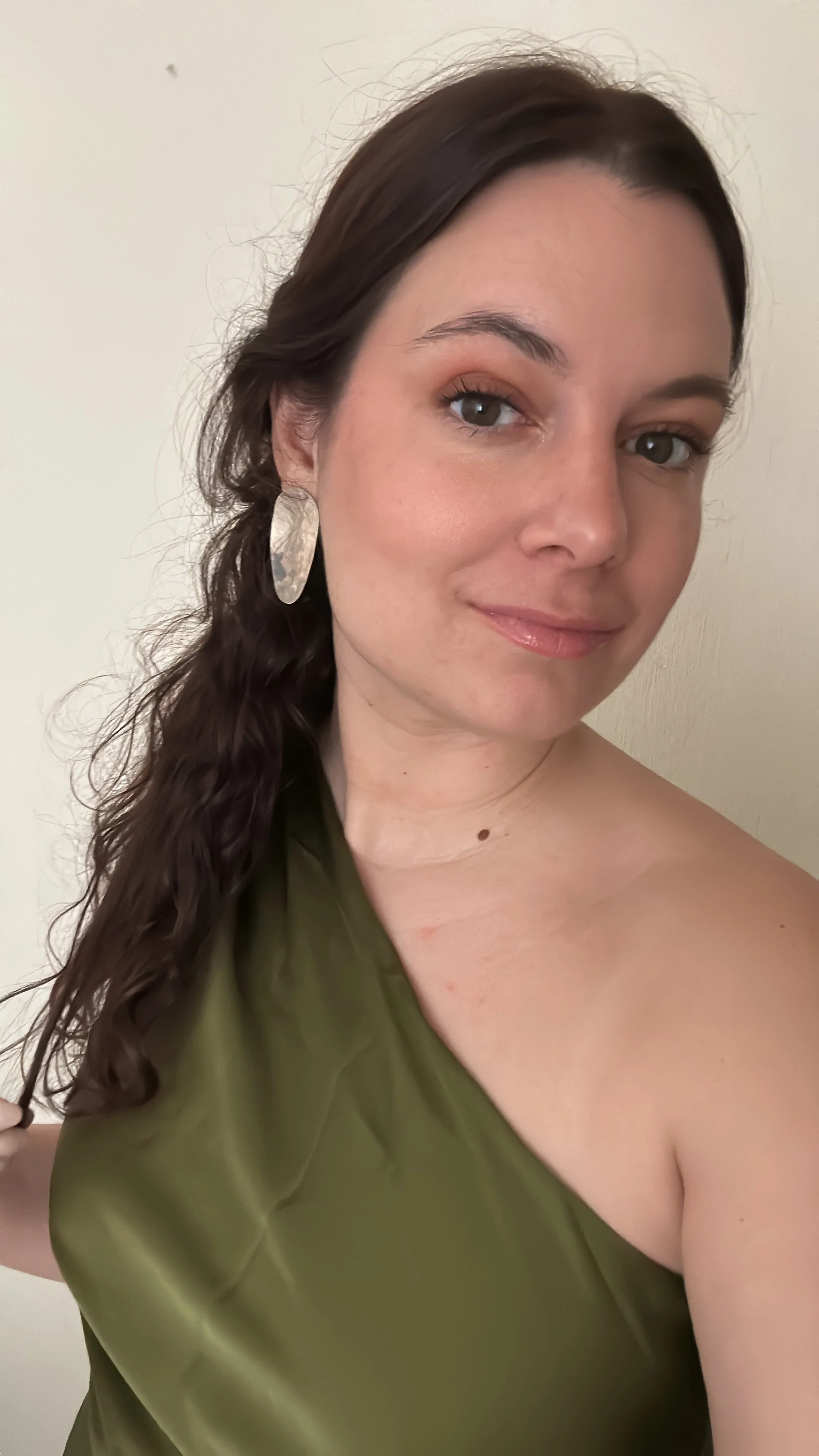 A woman with dark brown, wavy hair styled to one side, wearing a green, off-the-shoulder top, and large, oval earrings, taking a selfie against a plain light-colored wall.