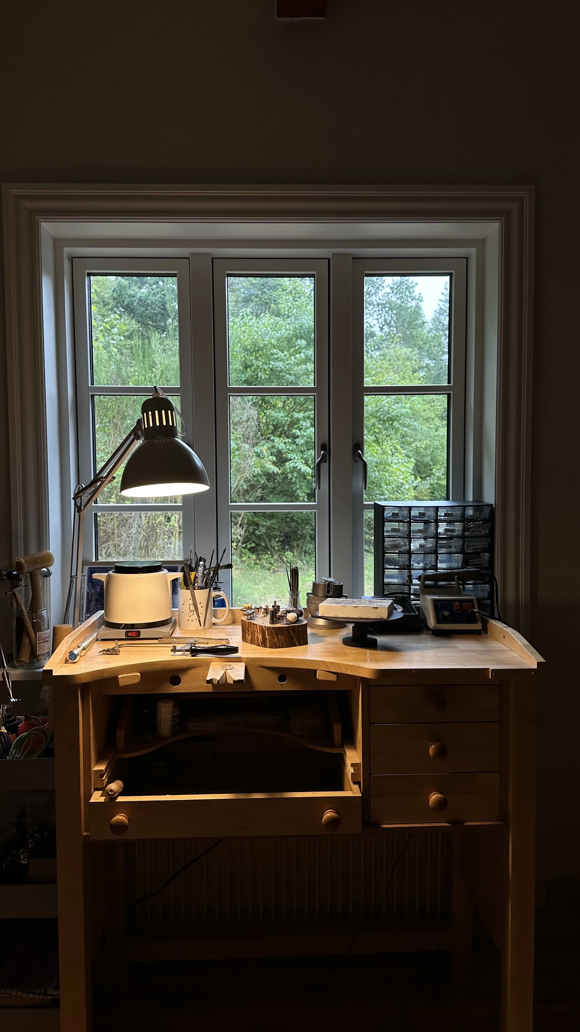 Workstation with a wooden desk near a window showing green trees outside, cluttered with tools, materials, and a focused task light.