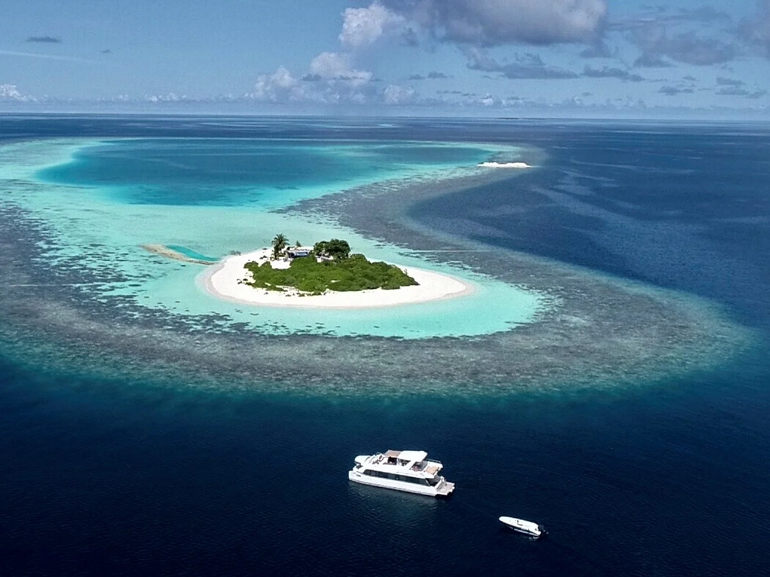 A small tropical island with white sandy beaches, lush green vegetation, and several buildings, surrounded by turquoise waters with coral reefs and a boat nearby in the ocean.