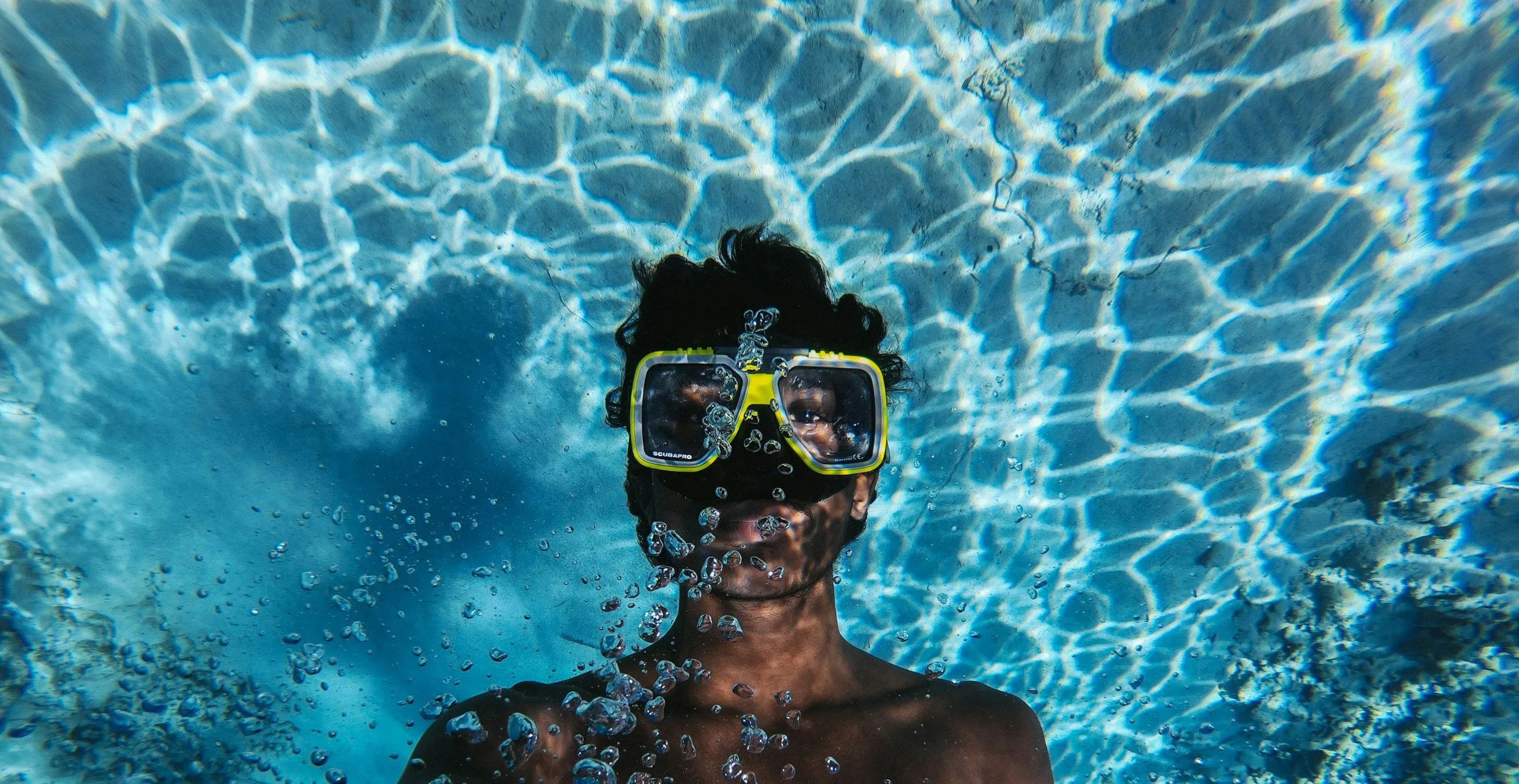 Person underwater wearing diving mask and snorkel in a swimming pool with rippling light reflections.