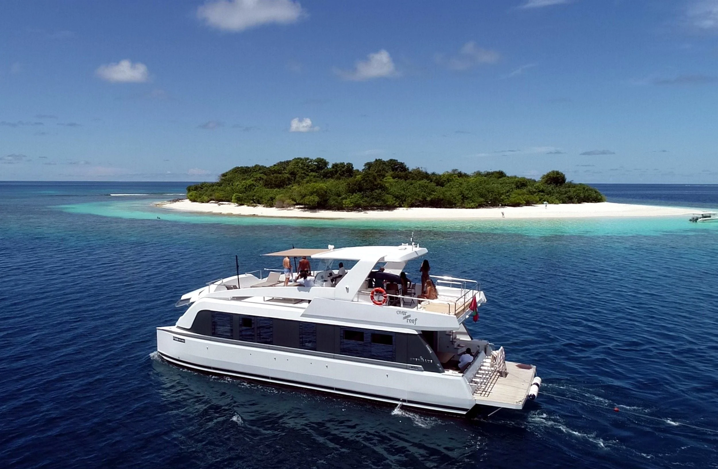 A white yacht sailing in clear blue water near a tropical island with white sandy beach and green trees, under a partly cloudy sky.