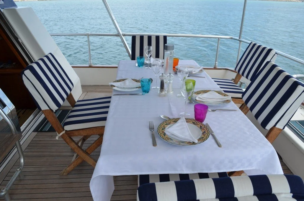 A dining table set for a meal on a boat deck, with six chairs featuring blue and white striped cushions, overlooking the water.