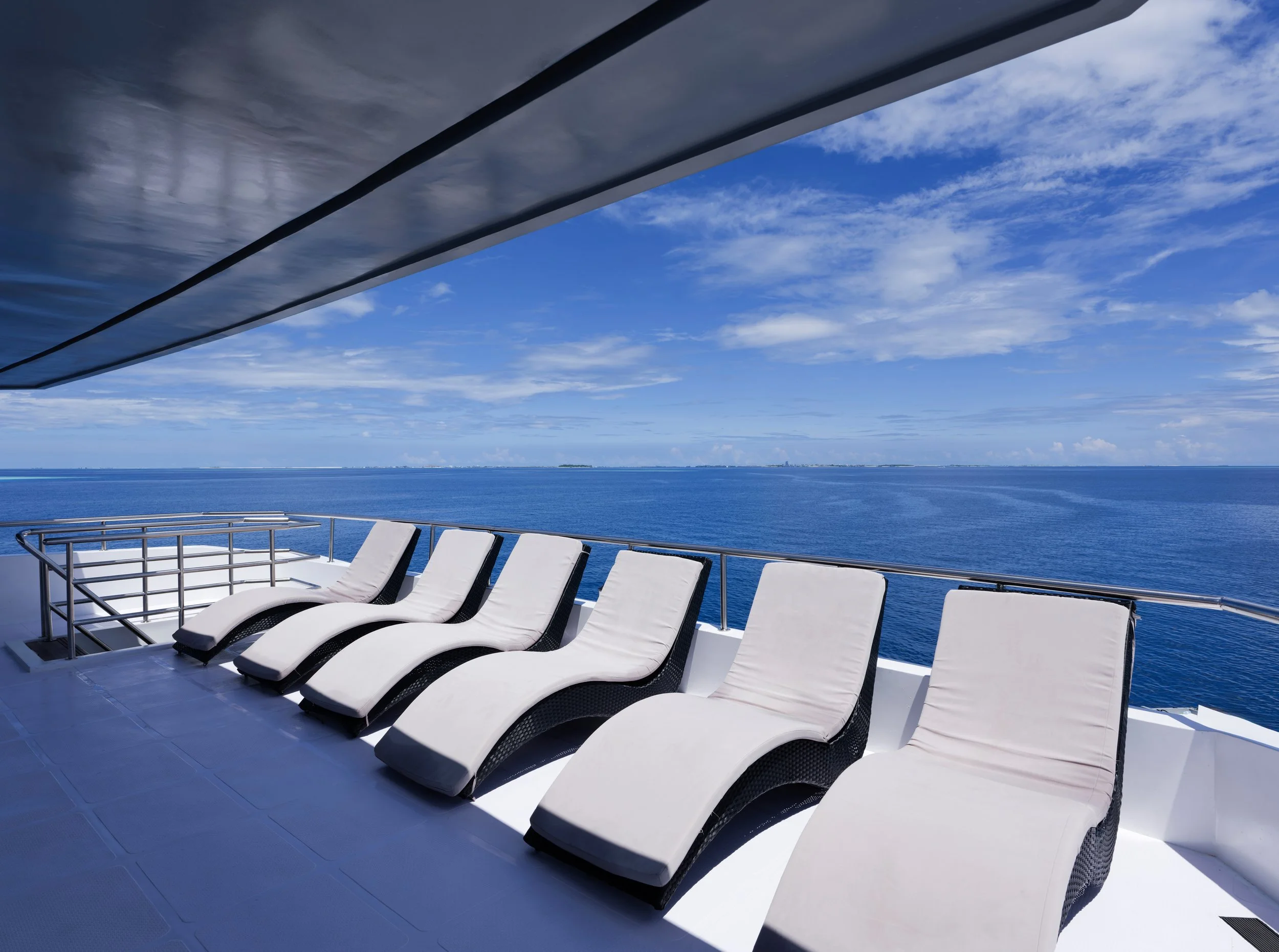 Seven white lounge chairs on a cruise ship balcony overlooking the ocean with a blue sky and scattered clouds.