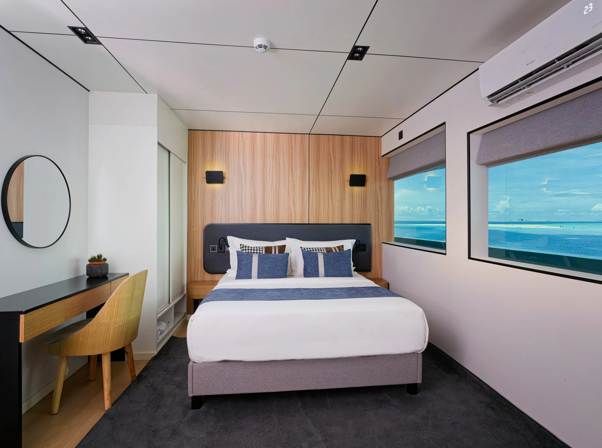 Modern hotel bedroom with a view of the ocean through large windows, a bed with dark and white bedding, a wooden desk, a mirror, and a small plant, in a minimalistic style.