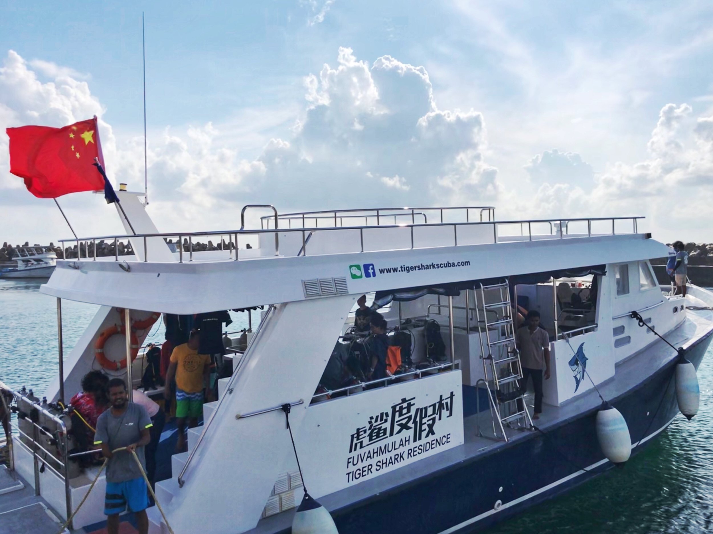 A white boat named 'Fuvahmulah Tiger Shark Residence' docked at a harbor with people on board. The boat has a red Chinese flag flying at the stern, and website and social media info on the side. The background shows a partly cloudy sky and other boat