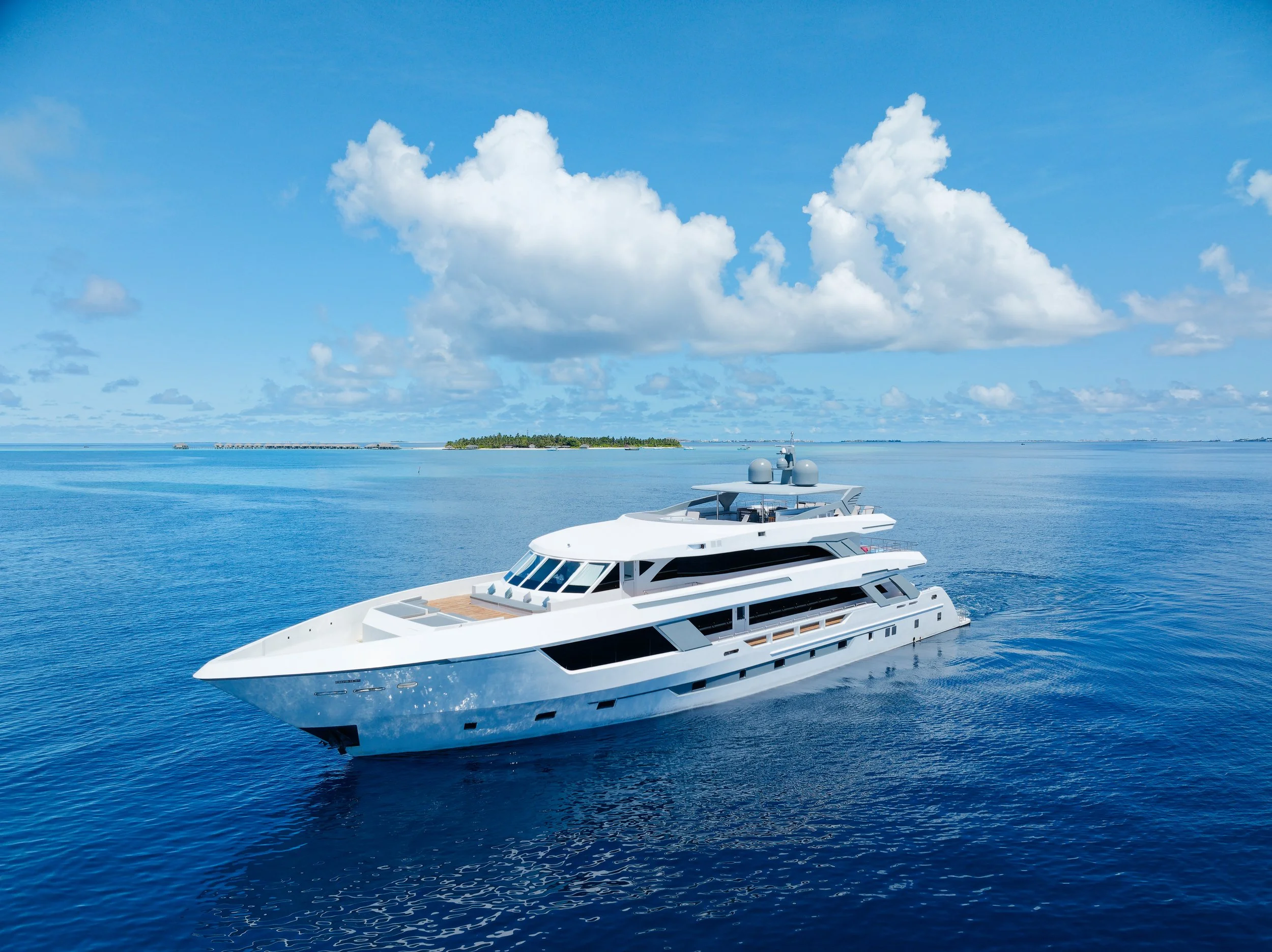 A large white luxury yacht sailing on calm blue ocean waters underneath a partly cloudy sky, with a small island in the background.