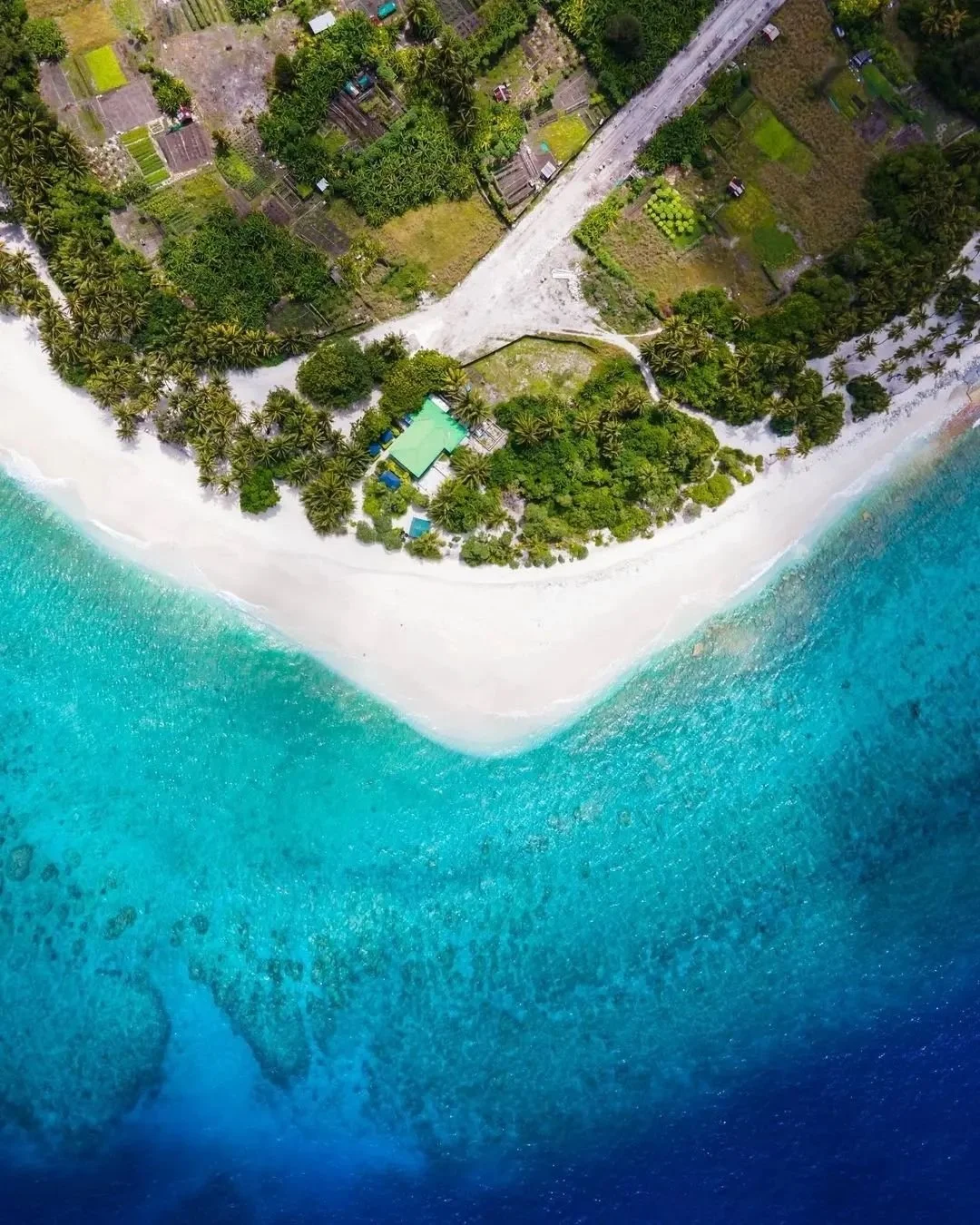 Aerial view of a tropical beach with white sand, turquoise water, lush green trees, and a small structure near the shoreline.