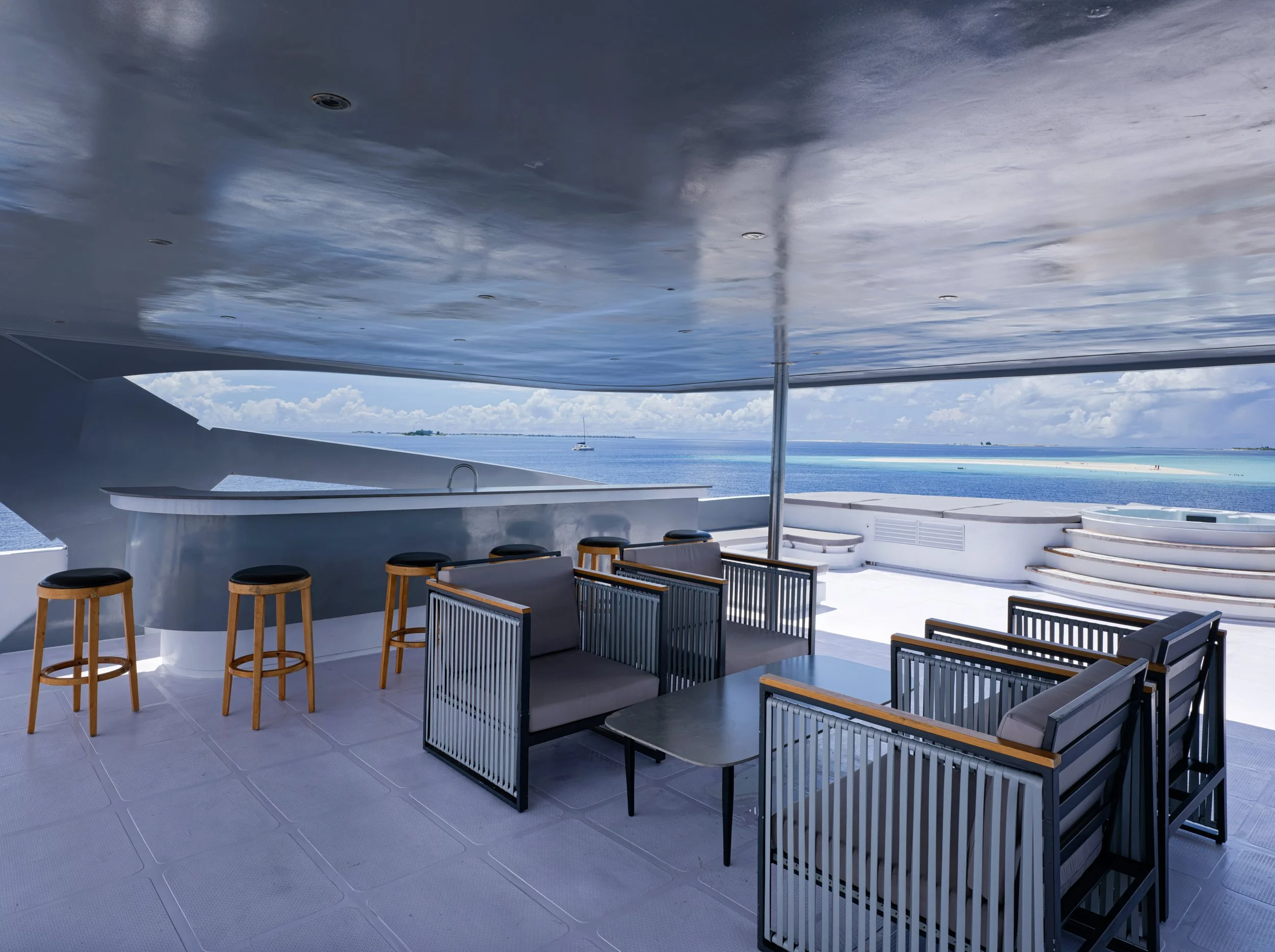 Outdoor seating area with gray and black chairs, a small table, bar stools, and a view of the ocean with a sailboat in the distance.