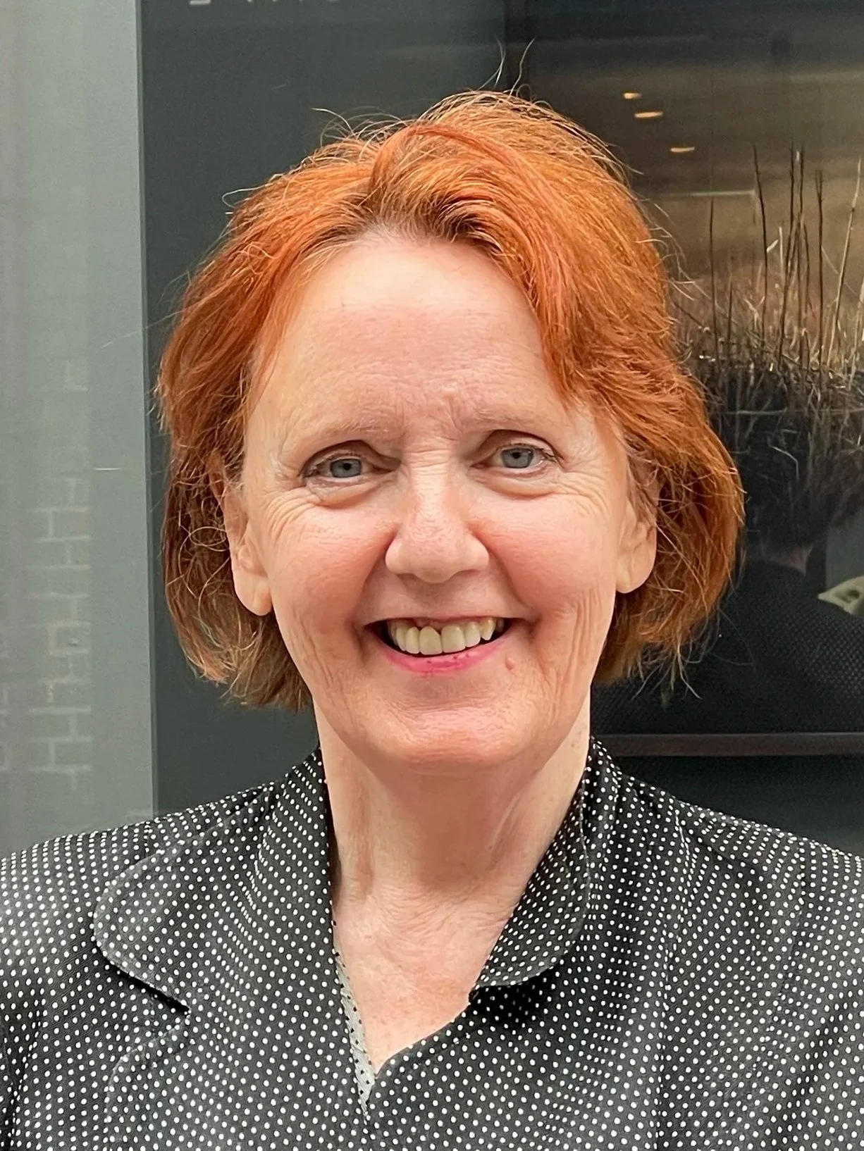 A woman with short brown hair, wearing sunglasses on her head, a green jacket with floral embroidery, and a red shirt, takes a selfie outdoors on a dirt path under a blue sky with sparse clouds, with yellow flowers and green shrubbery in the background.