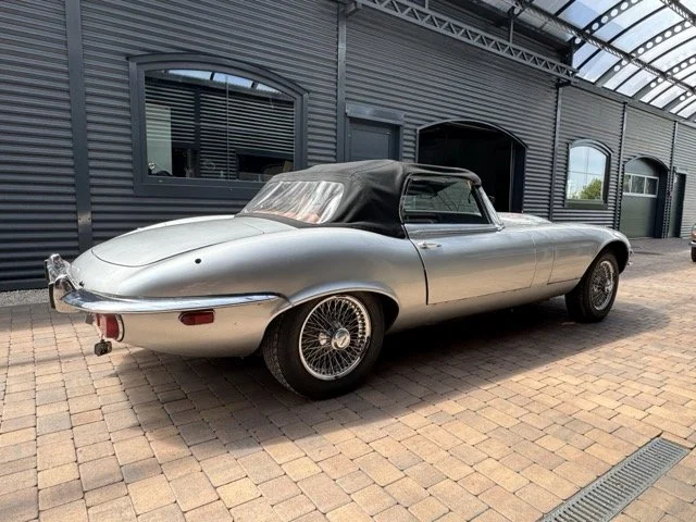 Silberner Jaguar Etype S3 V12-Cabriolet mit schwarz gepolstertem Dach, geparkt auf Kopfsteinpflaster vor einer schwarzen modernen Gebäudewand