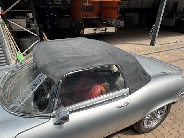 Ein silberner Oldtimer mit schwarzem Stoffverdeck, auf einem Hof mit Werkzeug und anderen Gegenständen im Hintergrund.