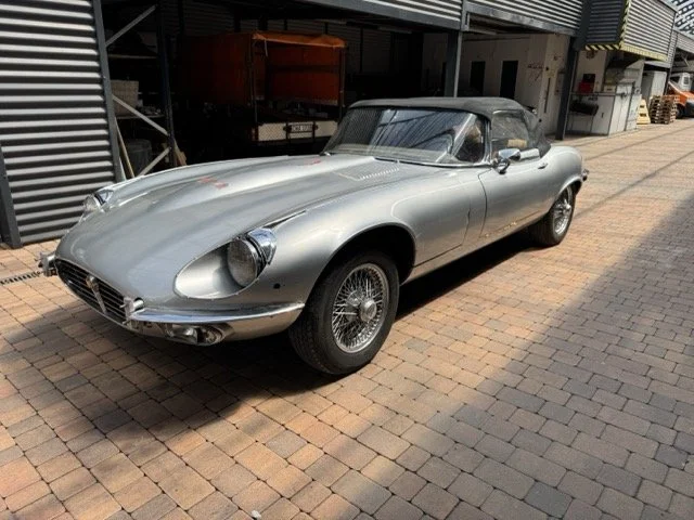 Ein silberner Jaguar Etype S3 V12 mit geschlossener Stoffdächer auf gepflastertem Boden in einer Werkstatt oder einem Außenbereich.