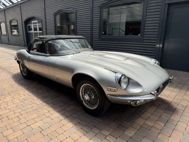 Ein silberner vintage Jaguar Etype S3 V12, parkend vor einem modernen Gebäude.