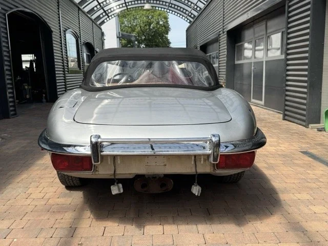 Ein silberner Jaguar Etype S3 V12 mit abnehmbarem Verdeck, geparkt unter einer gewölbten Metallkonstruktion auf gepflastertem Boden.