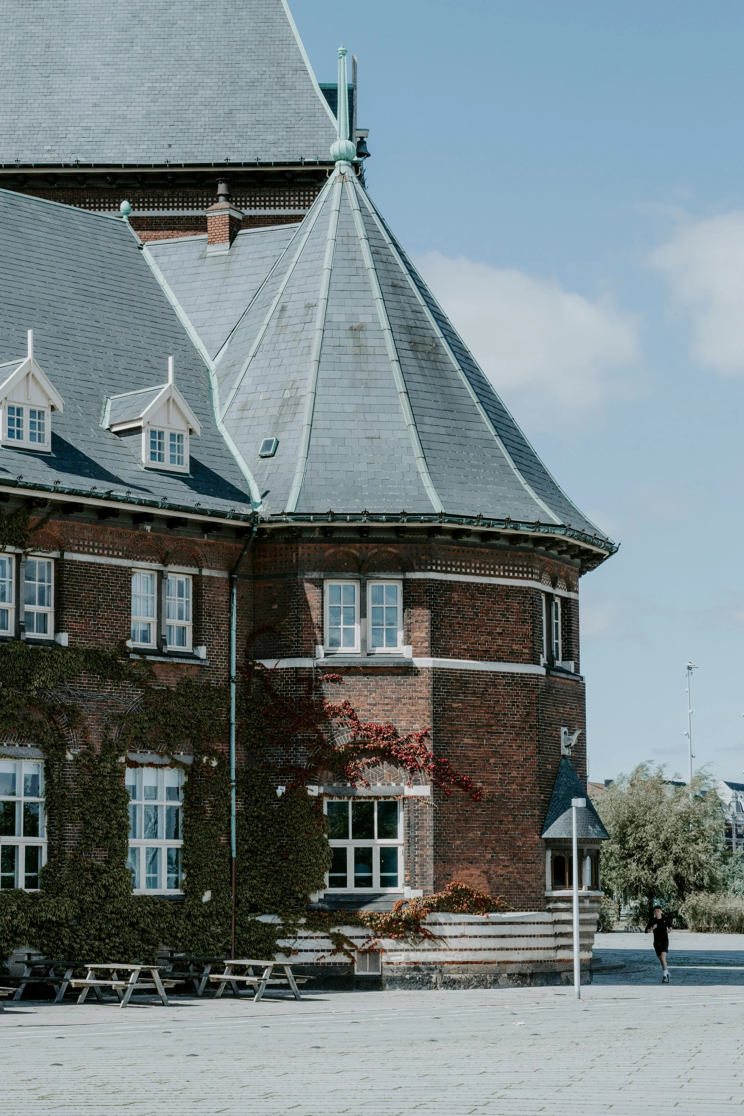 A historical brick building with a steep, gray slate roof and multiple small dormer windows, surrounded by an open paved area and some trees, under a partly cloudy sky.