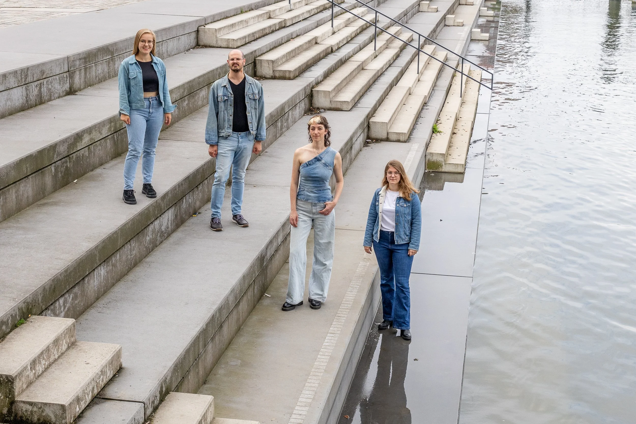 Four women standing on concrete steps by a body of water, dressed in casual denim outfits.
