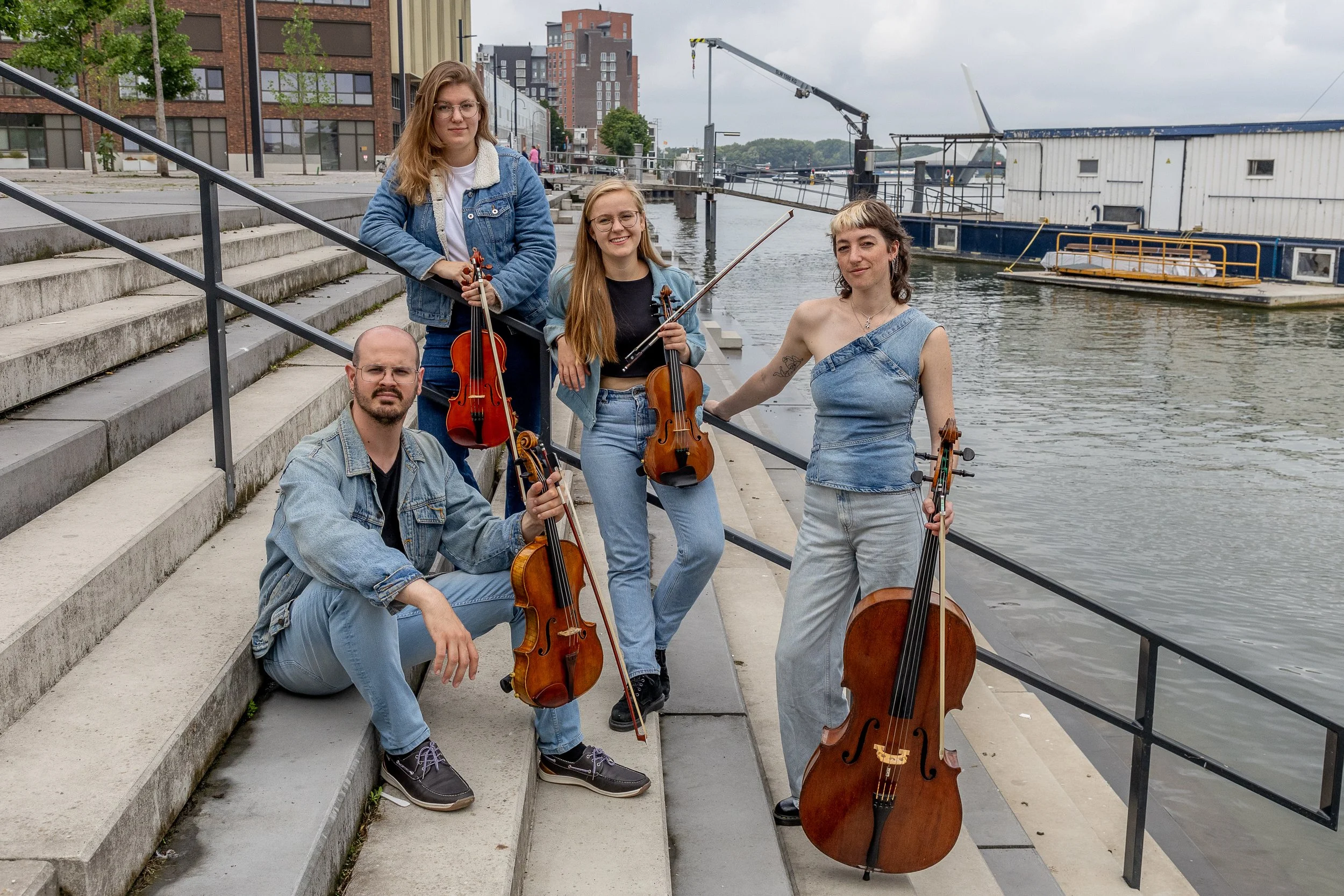 Group of five young musicians, holding string instruments, standing and sitting on concrete steps by a river, with apartment buildings and boats in the background.