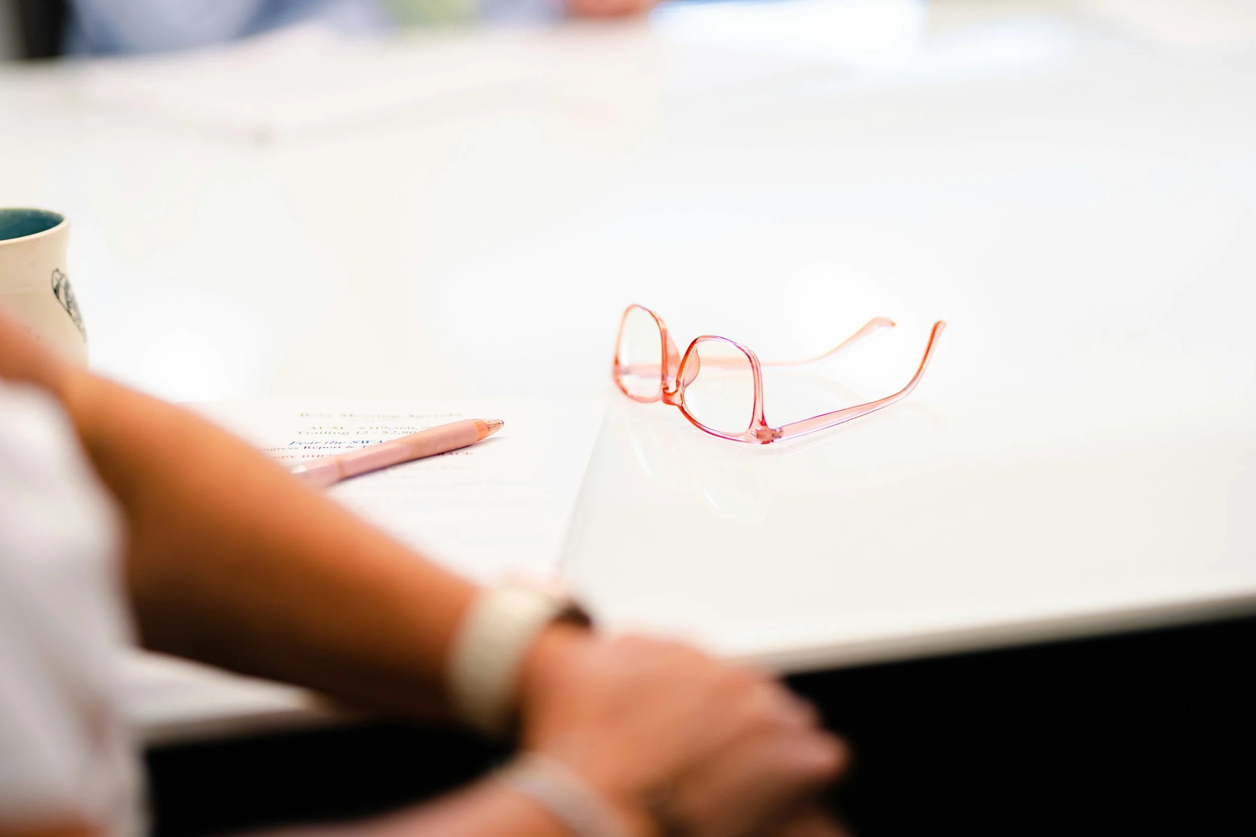 A pair of pink eyeglasses rests on a white table near a pink pen and a partially visible paper, with a person's arm and wristwatch also visible in the foreground.