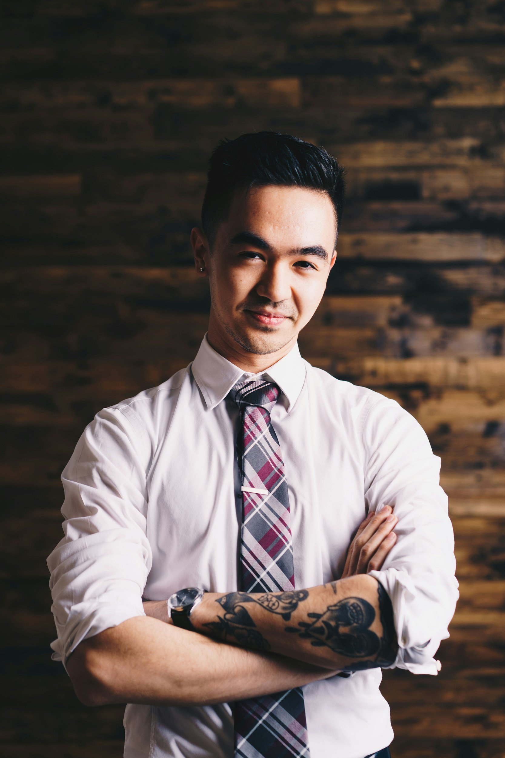 A confident man with tattoos on his left arm, wearing a white dress shirt and a plaid tie, stands with arms crossed in front of a wooden wall.