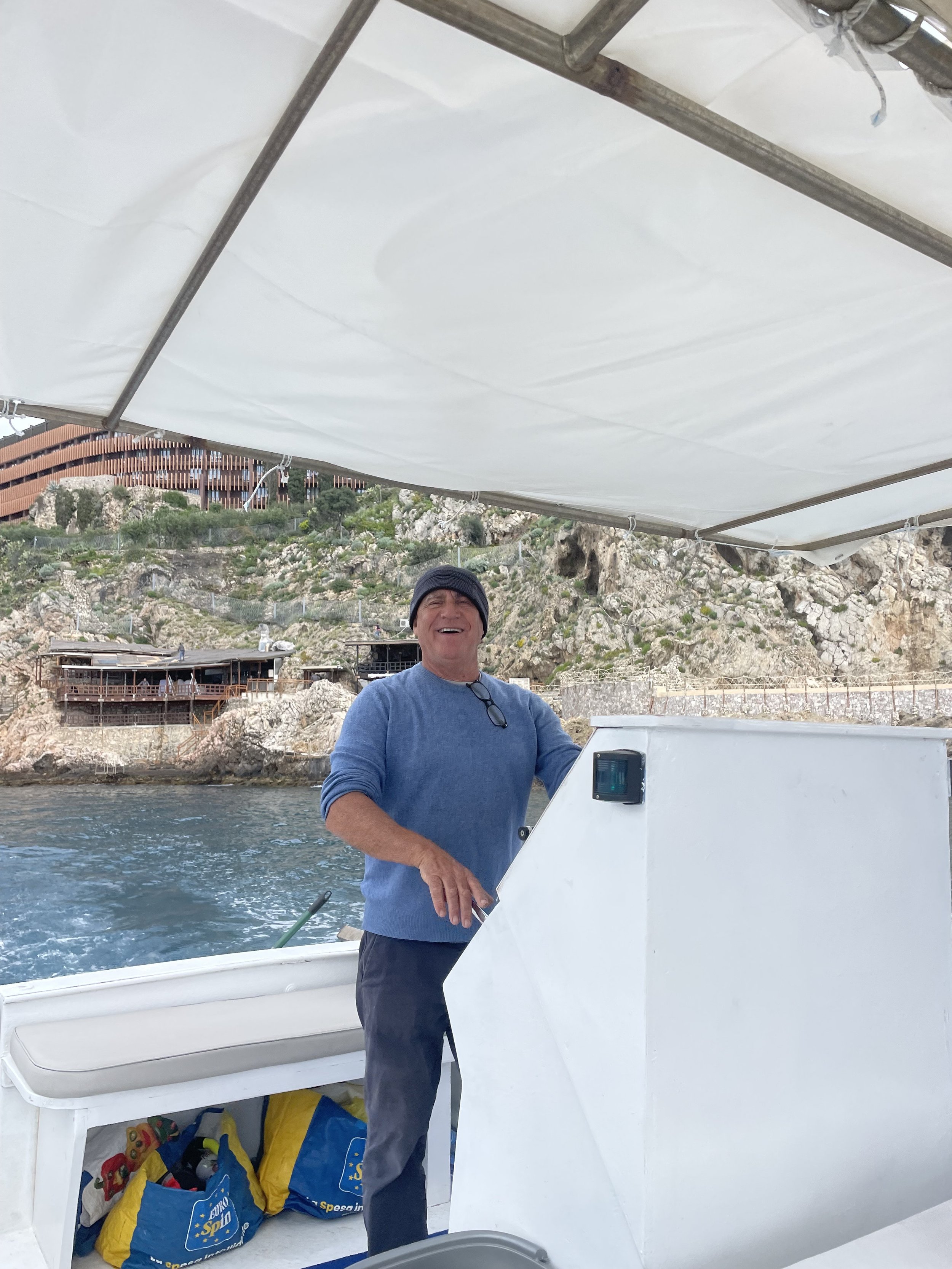 Person steering a boat near rocky coastline with a building in the background.