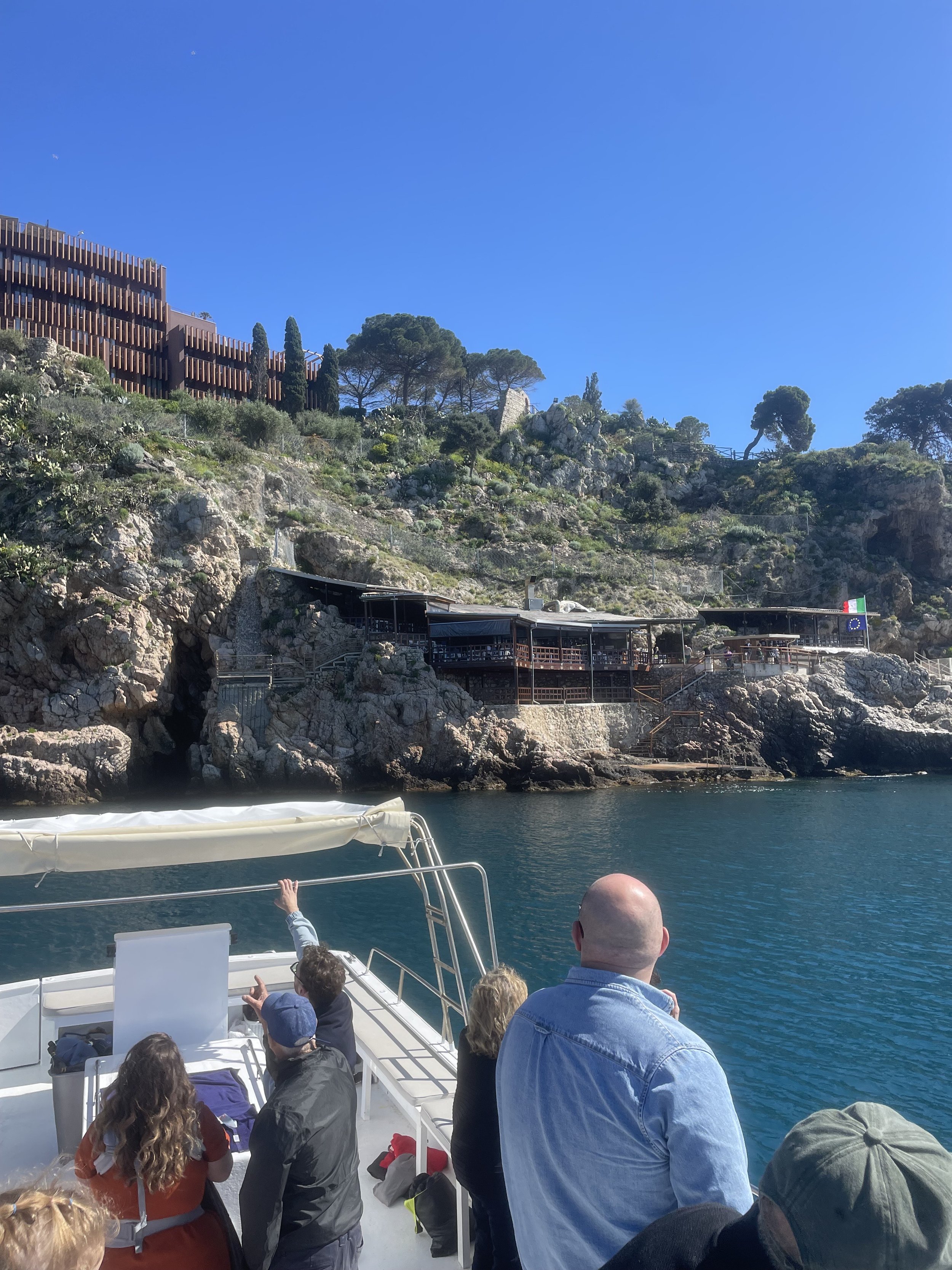 People on a boat observing a rocky coastline with a cliffside building and greenery.