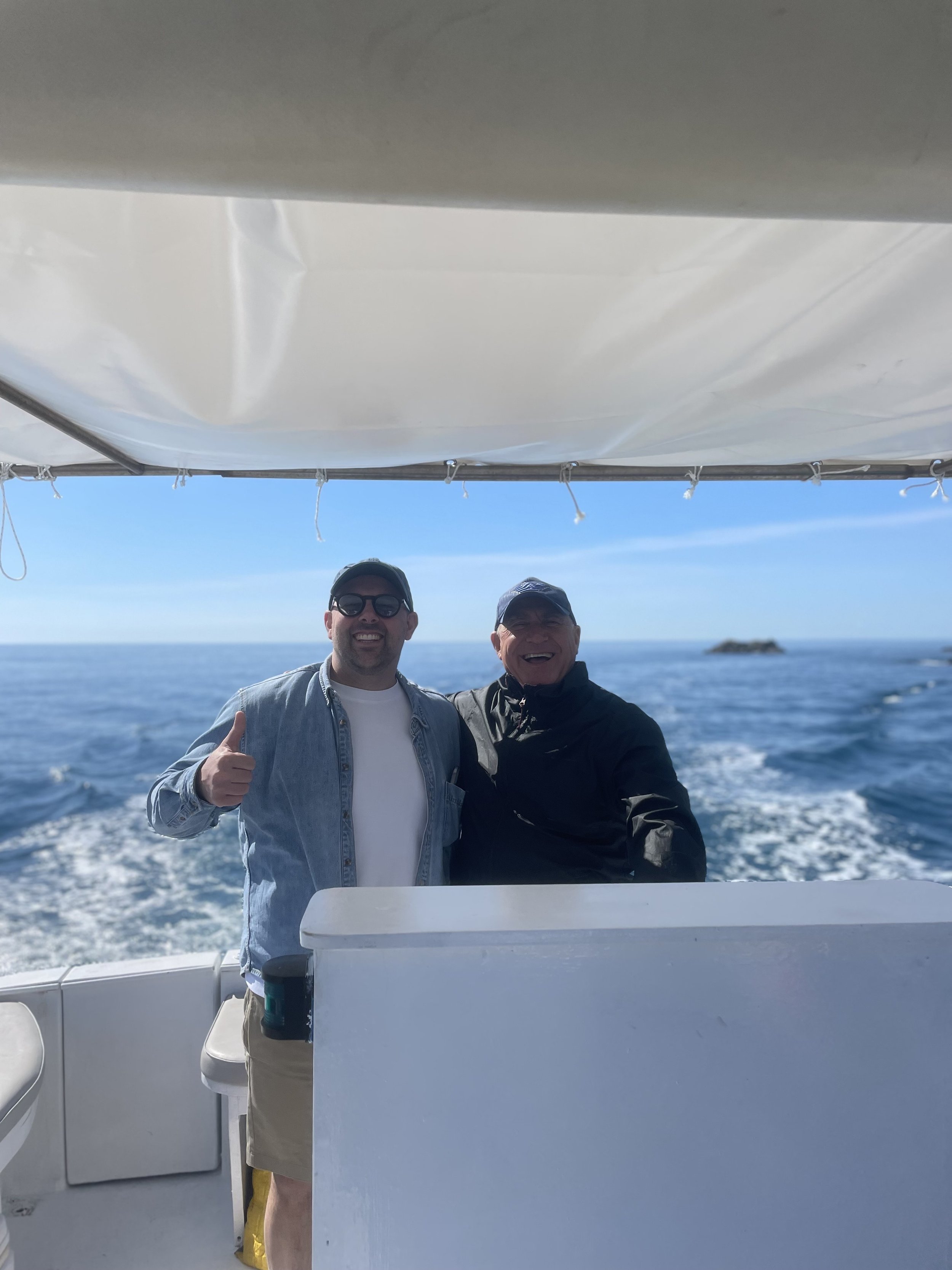 Two men smiling on a boat in the ocean, one giving a thumbs-up, with a blue sky in the background.