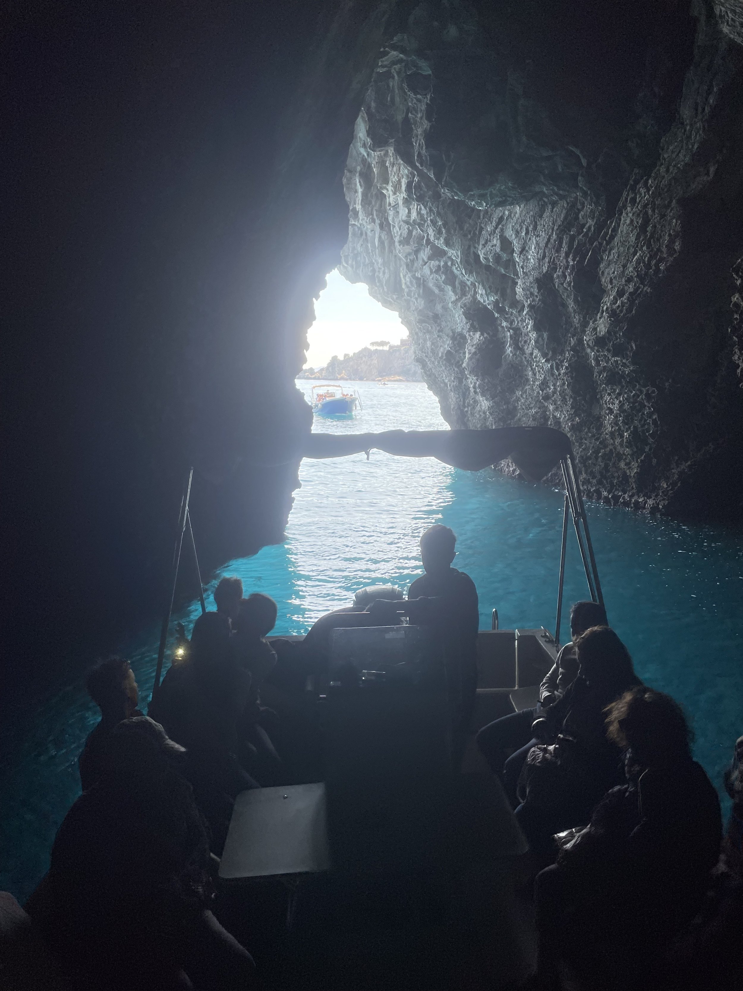 People on a boat inside a cave, with blue water and an opening leading to the outside.