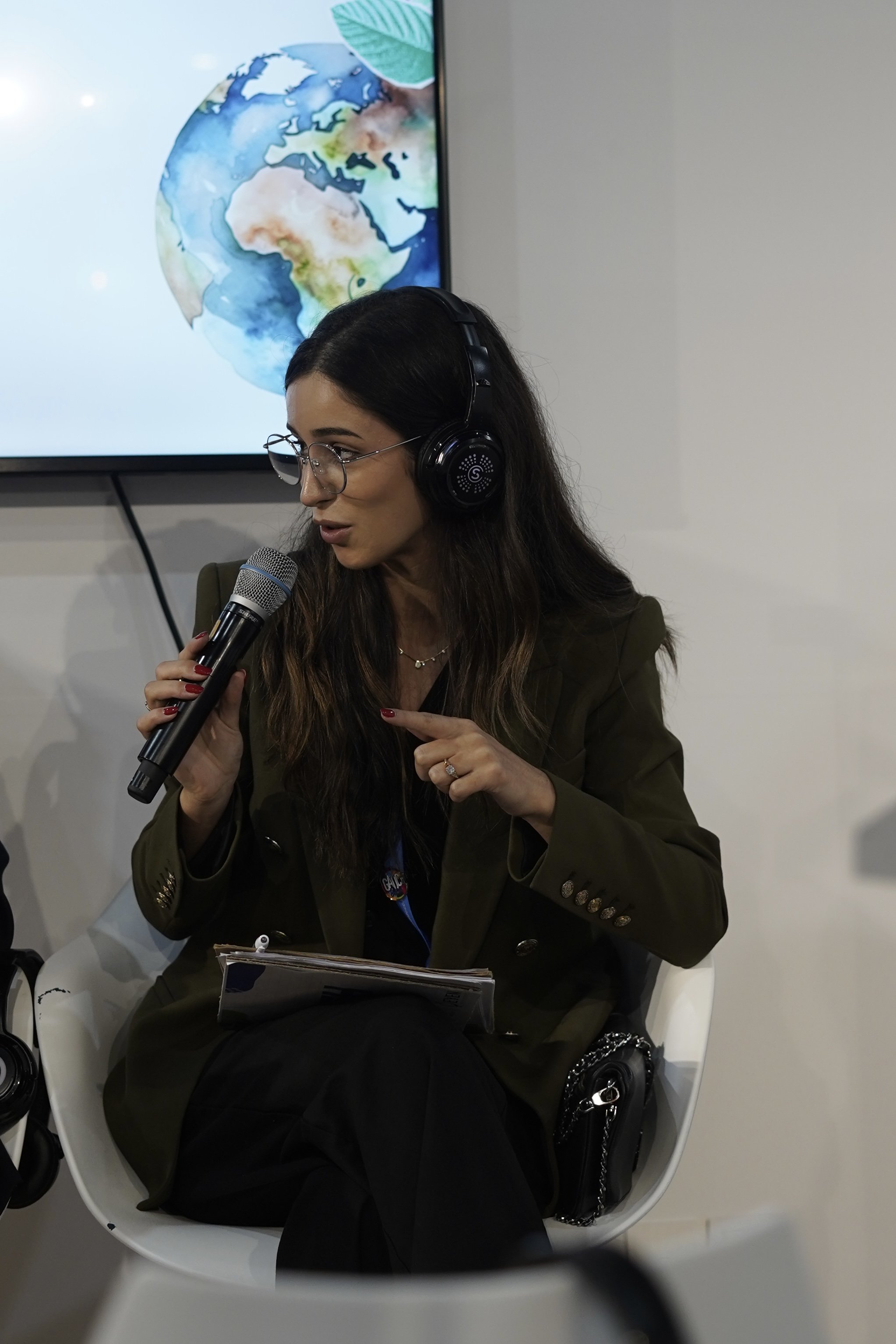 A woman sitting on a white chair, speaking into a microphone, wearing headphones and glasses, with a notebook on her lap. A television screen behind her displays a watercolor map of the world.
