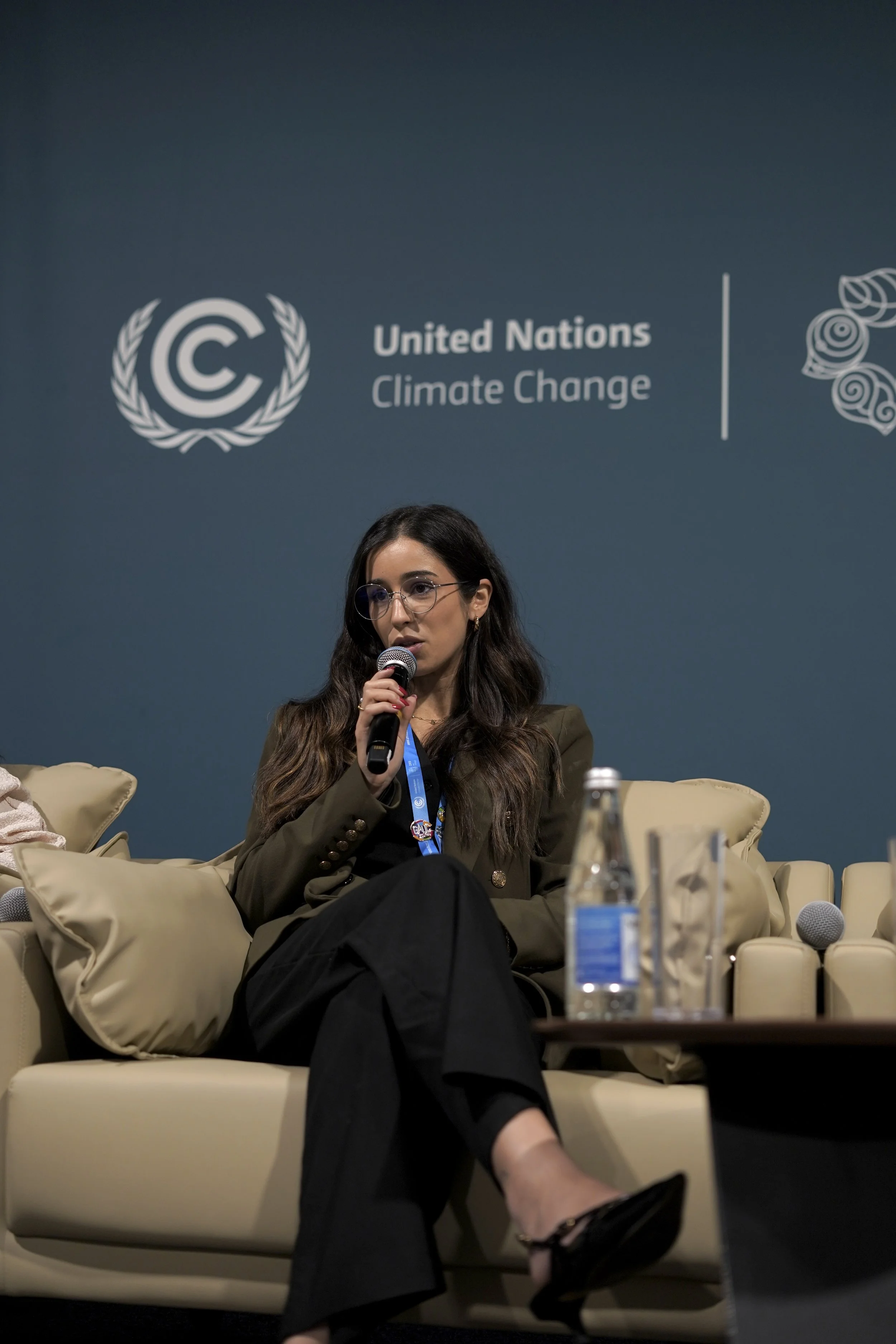 A woman is sitting on a beige couch at a United Nations Climate Change event, holding a microphone. She has long wavy hair, glasses, and is wearing a dark blazer. There is a table with bottled water and glasses in front of her. The background features the United Nations Climate Change logo and text.