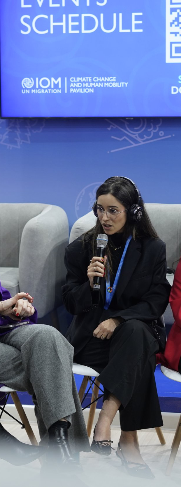A woman with long dark hair, glasses, and a black outfit speaking into a microphone at a panel discussion about climate change and human mobility at the UN Migration event.