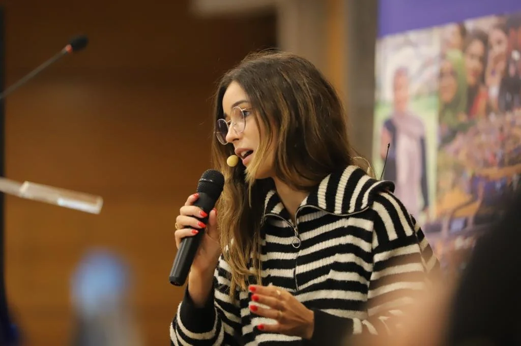 A woman with long brown hair and glasses speaking into a microphone at an indoor event, wearing a black and white striped hoodie.