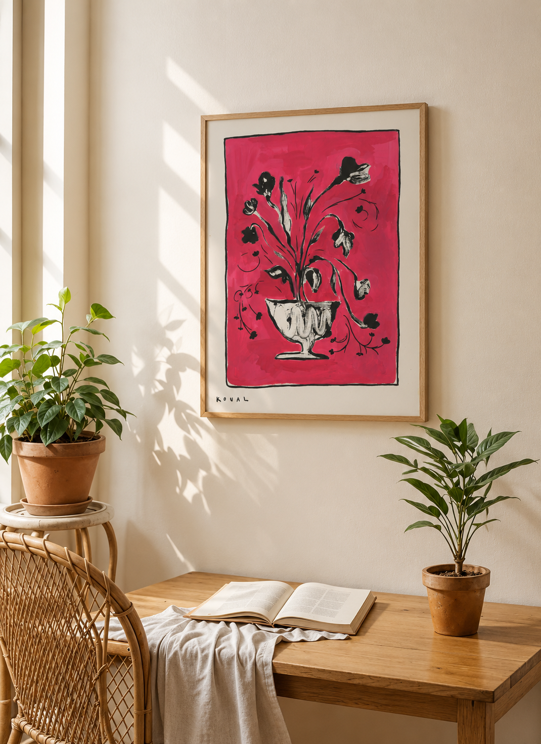 Framed print “Pink Flowers in a Vase” on a wall above a wooden desk with an open book, a rattan chair and two potted plants