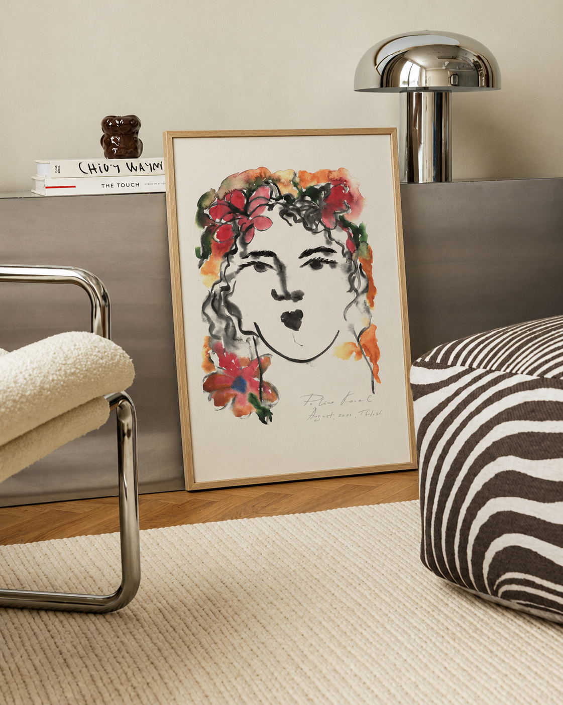 Framed print “August Girl” on a shelf next to a lamp and a modern chair with a striped cushion in a neutral room