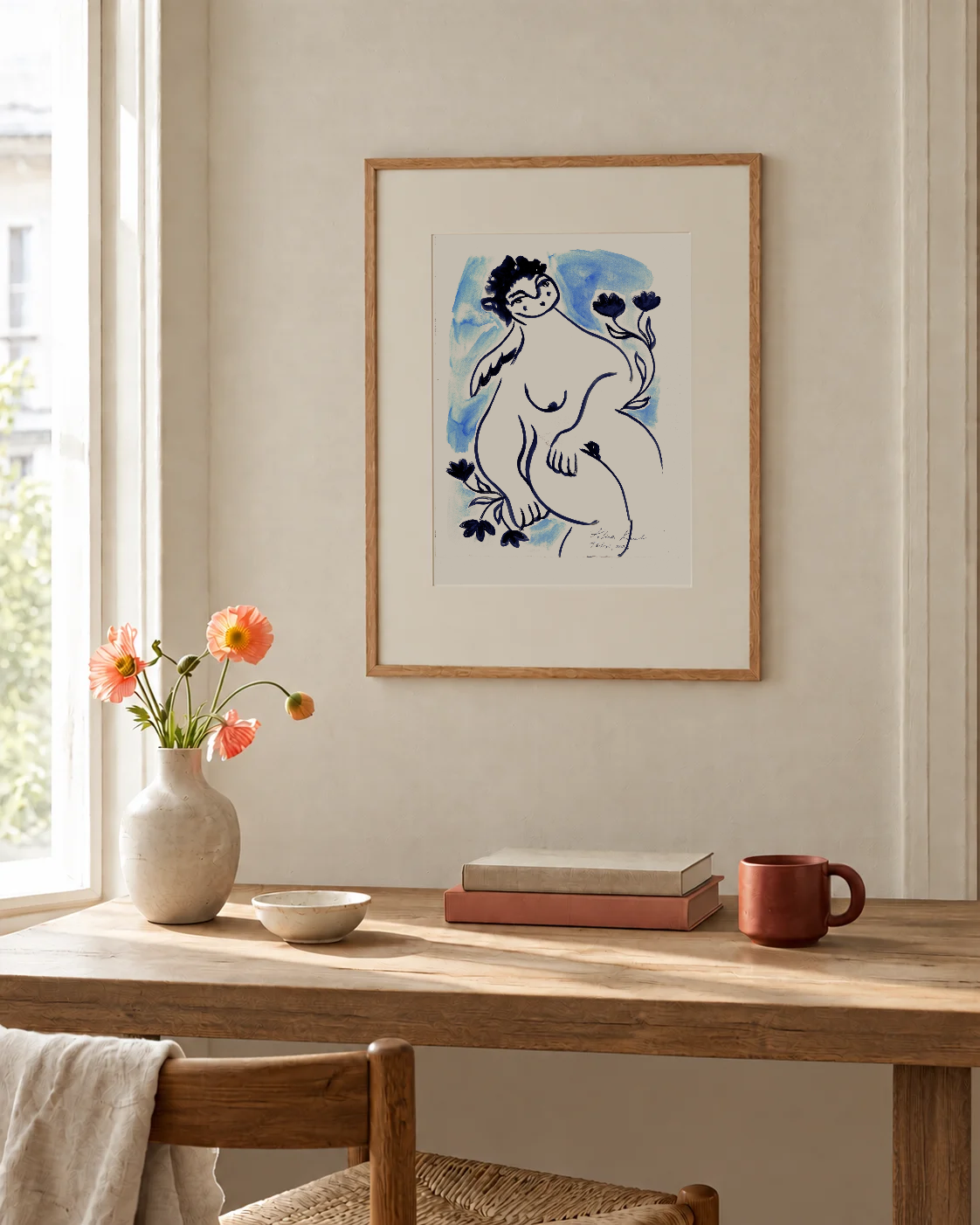 Angel by the Seaside framed print in an oak frame with a white mat, hanging above a wooden desk with a vase of poppies in a sunlit room.