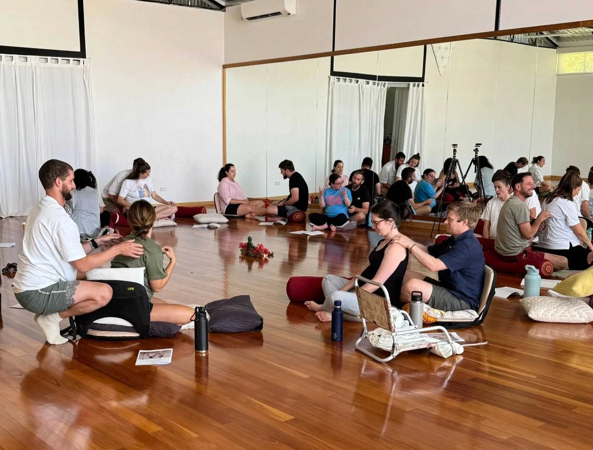 Meditation or yoga class in a spacious, well-lit studio with wooden floors and large mirrors. Participants are seated on cushions arranged in a circle, some receiving gentle neck or shoulder massages from facilitators. The atmosphere appears calm and focused.