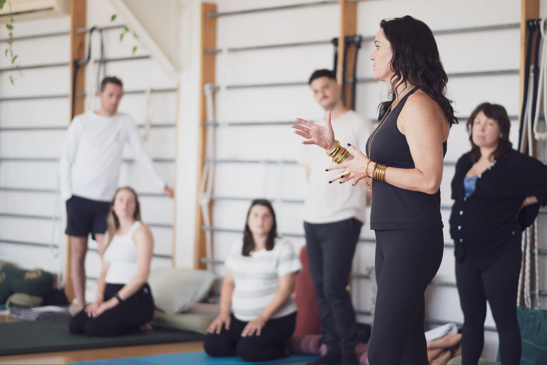 Woman in black speaking to a group during a fitness or yoga class in a gym studio, with people sitting and listening on a mat.