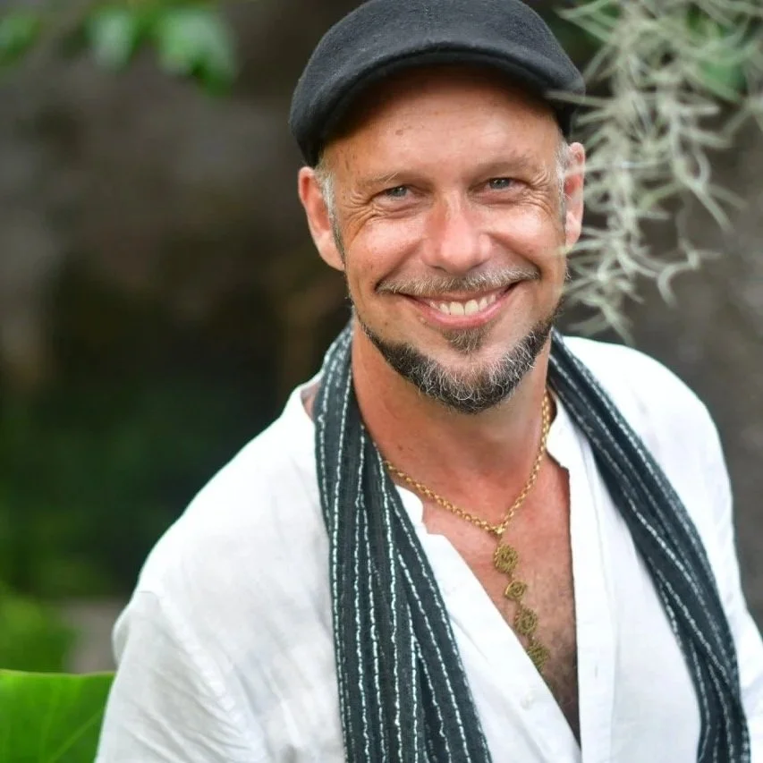 A smiling man with a beard and mustache wearing a black beret, white shirt, black and white striped scarf, and a gold necklace, standing outdoors with blurred greenery in the background.