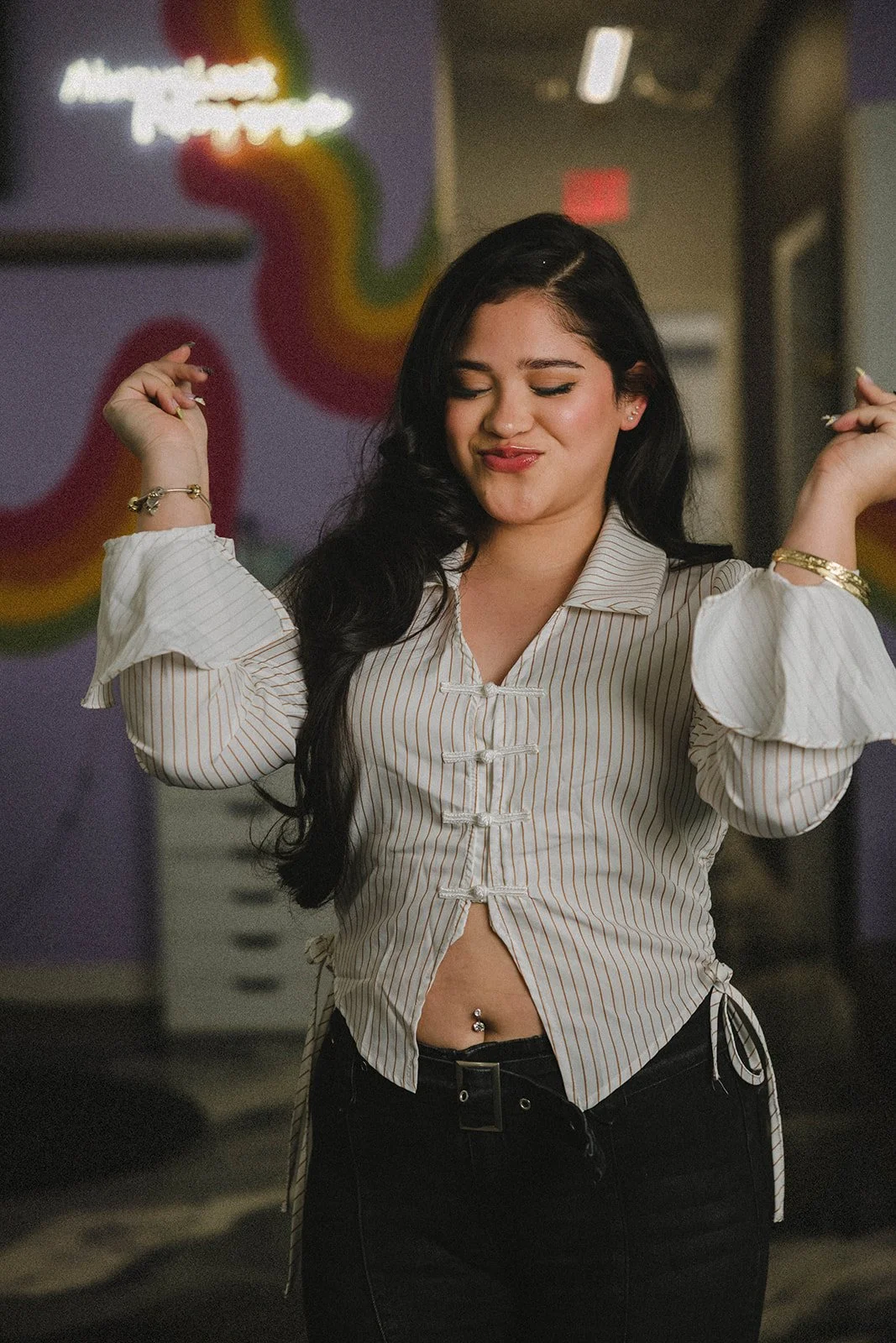 Daniela  with long dark hair and a belly piercing is dancing with her eyes closed and a smirk on her face. She is wearing a white striped blouse with puffed sleeves and black pants. The background features a colorful rainbow mural and neon lights.
