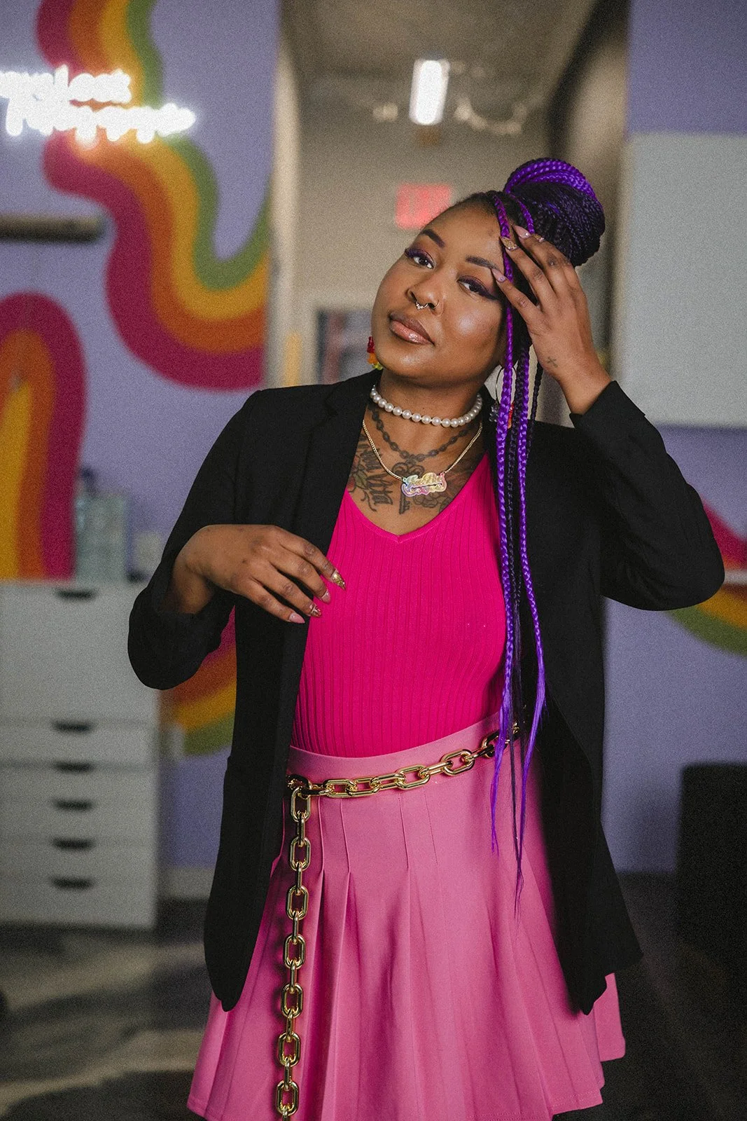 A woman with purple braids wearing a black blazer over a pink top and a pink skirt, accessorized with a pearl necklace, layered chains, and a gold chain belt, standing in front of a wall with colorful rainbow designs.