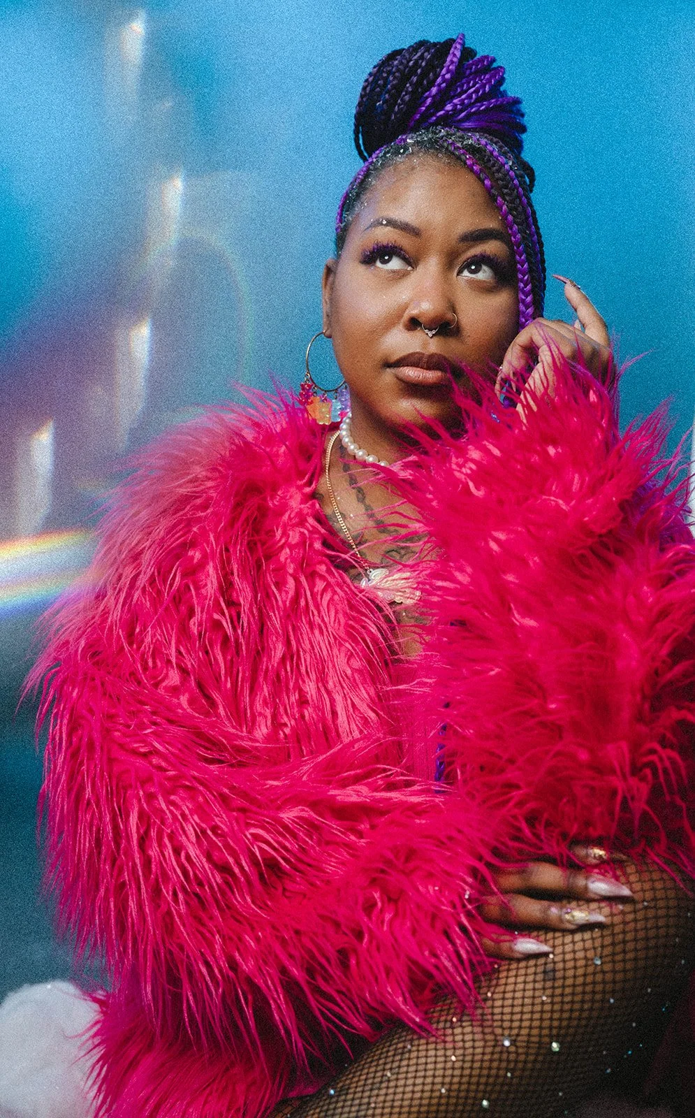 A woman with braided purple and black hair styled in a high bun, wearing a bright pink feathered jacket, fishnet stockings with rhinestones, jewelry including necklaces and hoop earrings, and standing against a blue background with rainbow reflections.