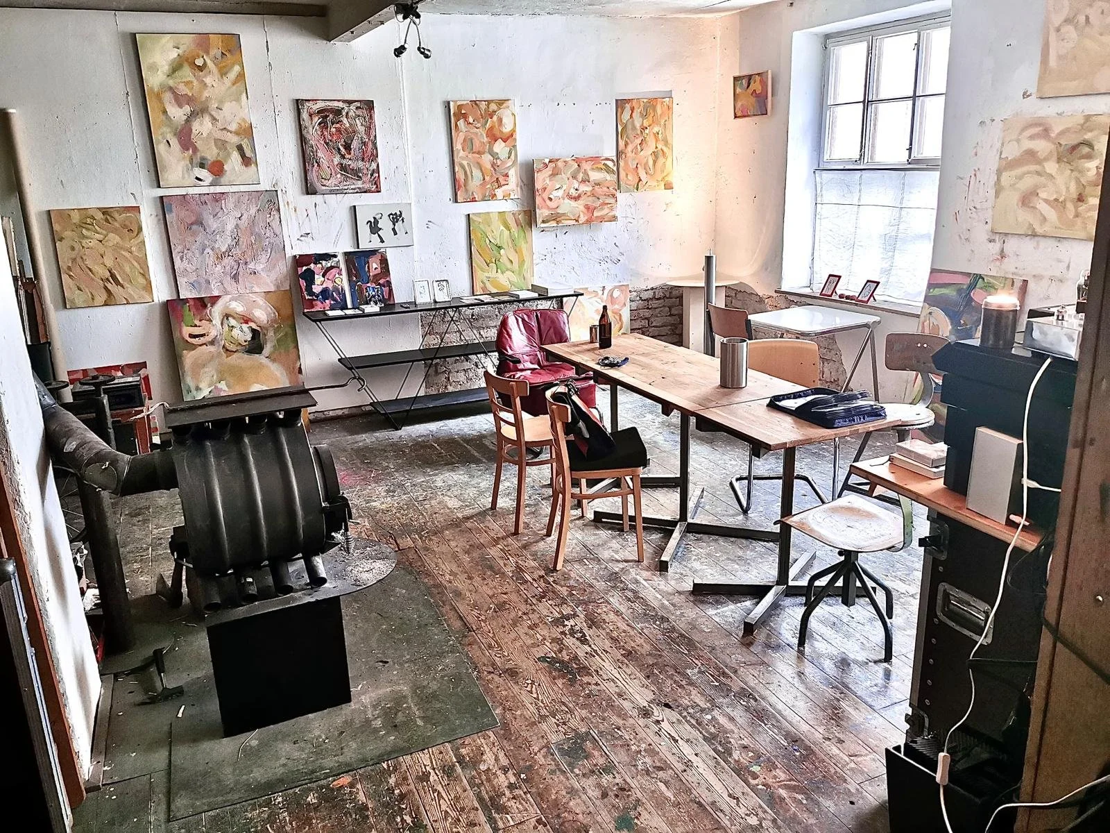 An artist's studio with abstract paintings on the walls, a wooden table with chairs, and art supplies. Sunlight streams through a large window.