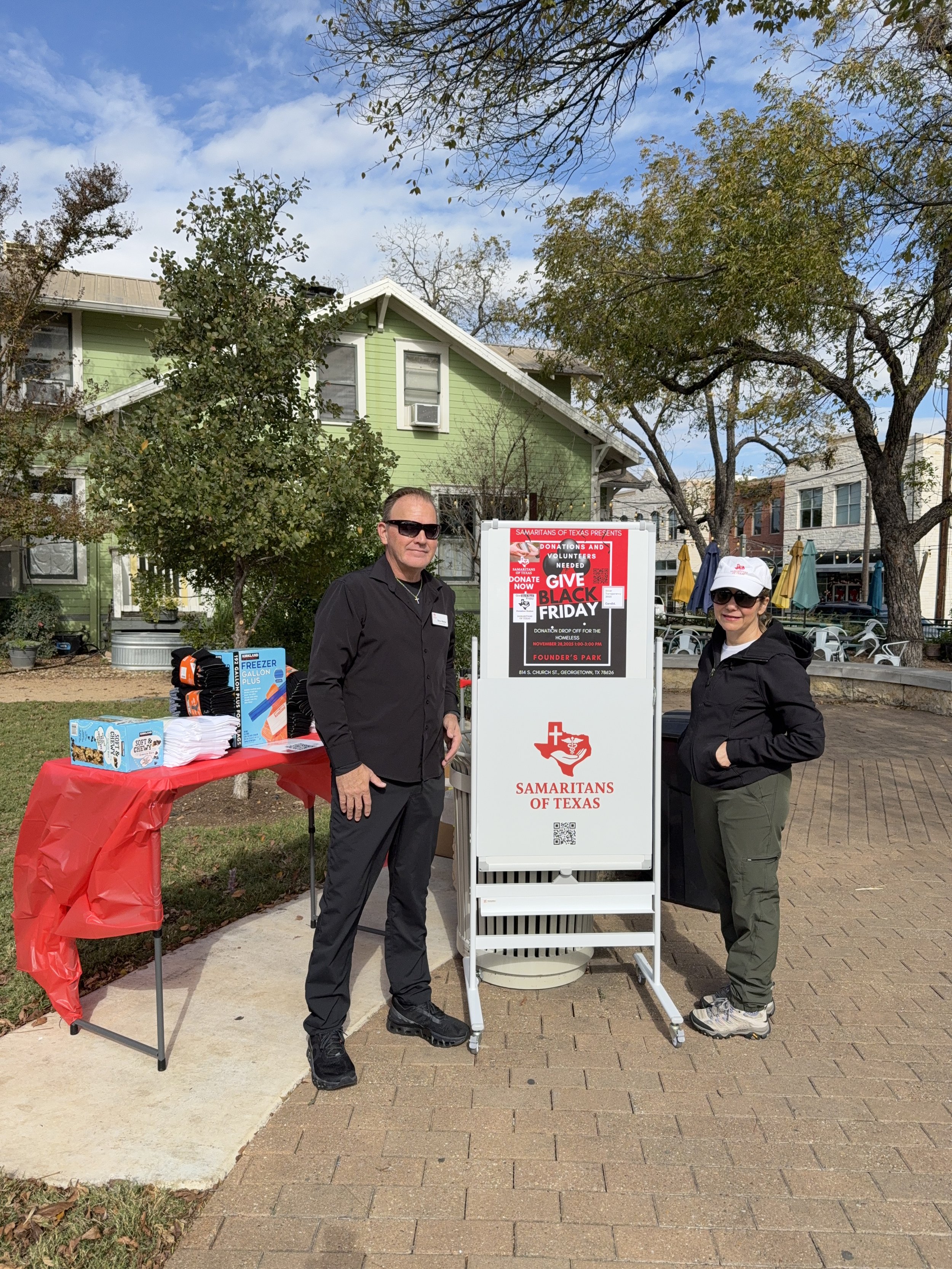 Two volunteers standing outdoors next to a sign for Samaritans of Texas, promoting Black Friday donation drive. A table with donation supplies is on the left, and there are trees and a green house in the background.
