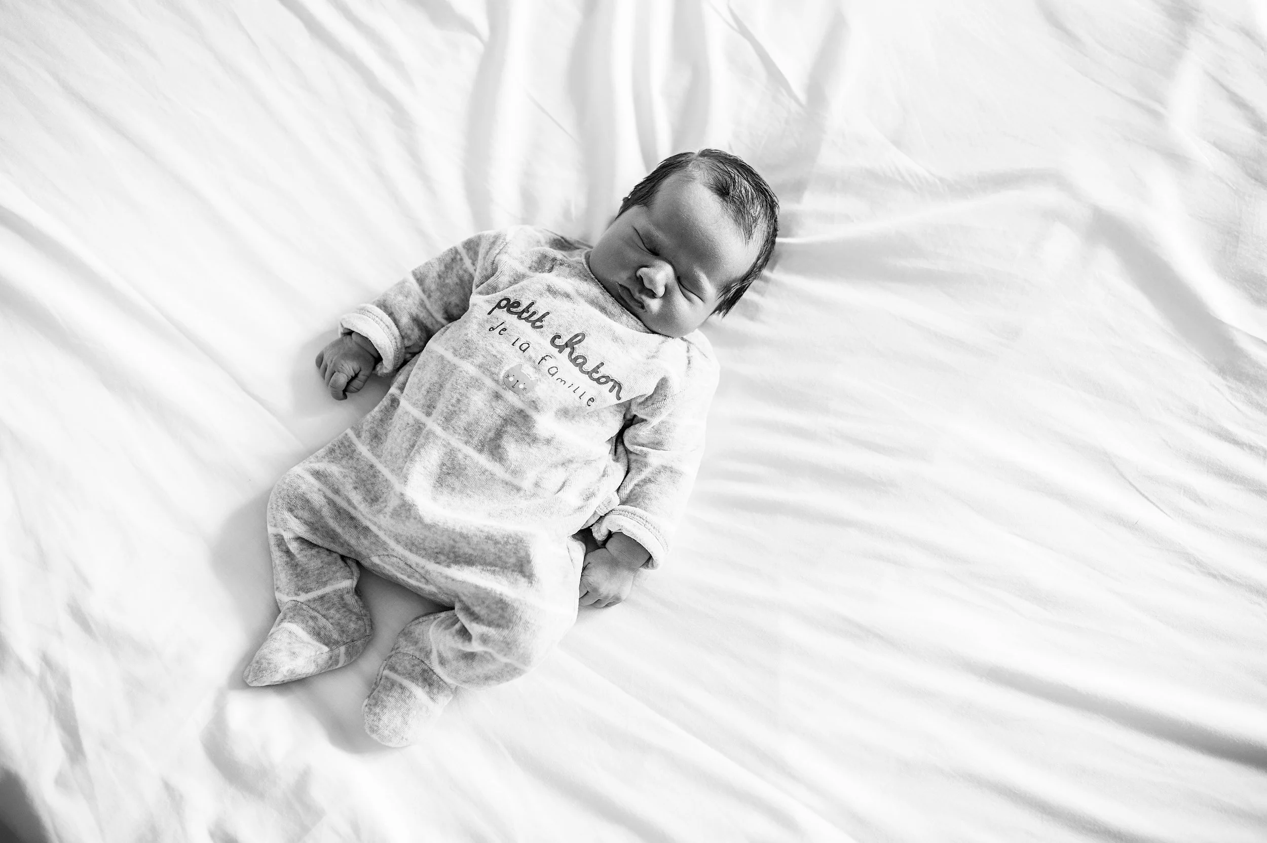 Un bébé endormi sur un lit blanc, portant un pyjama avec des mots français et un motif de rayures.