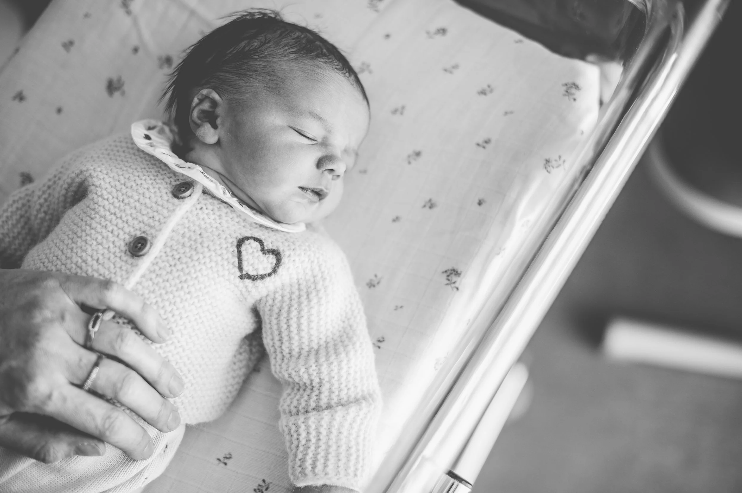 Un bébé endormi dans un lit d'hôpital, vu en gros plan en noir et blanc, avec une main d'adulte dans le cadre, probablement celle d'une infirmière ou d'un parent.