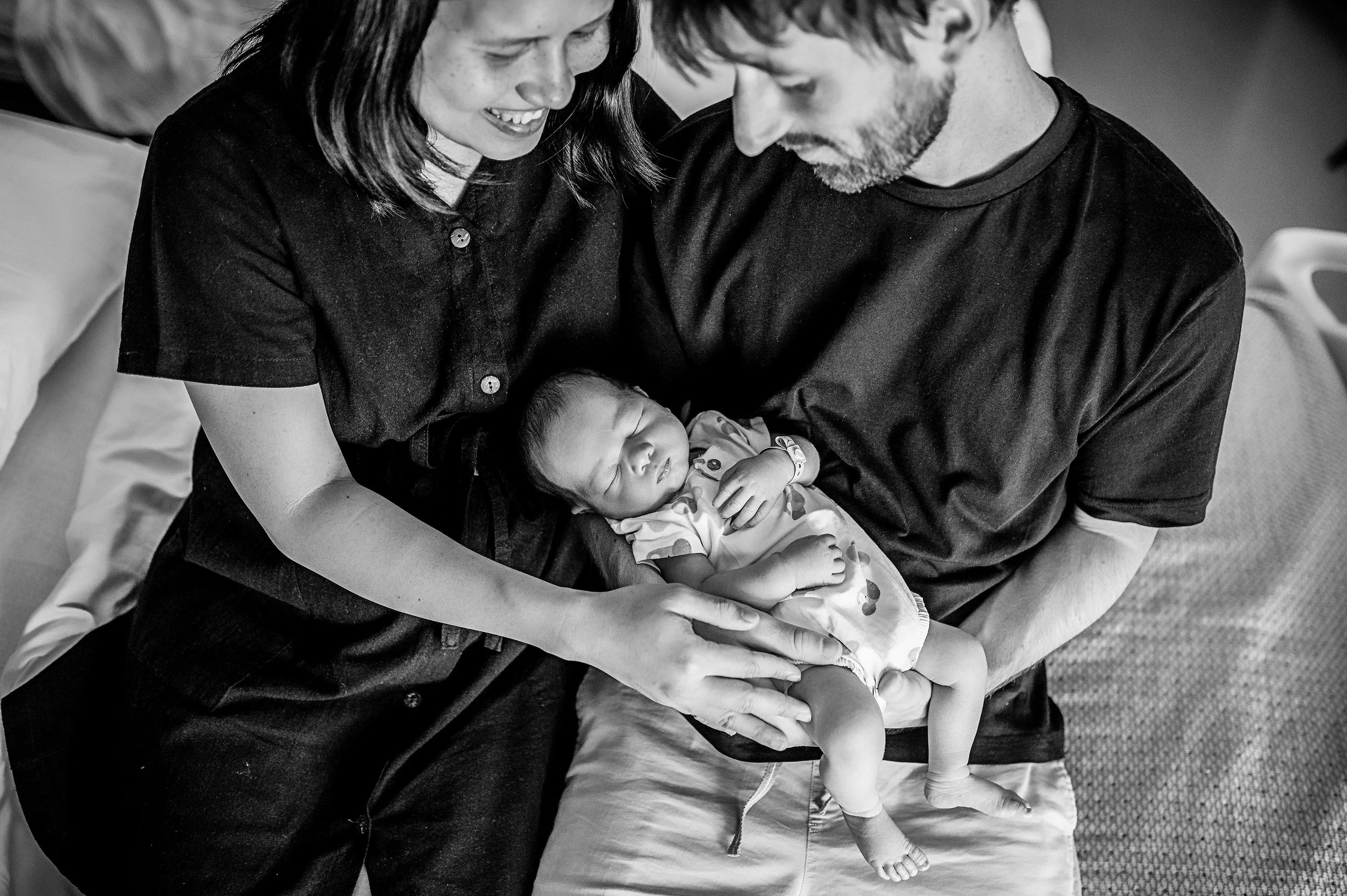 Une famille avec un nouveau-né dans une salle d'hôpital, la mère en noir, le père en noir, qui sourient et regardent l'enfant qui dort dans les bras du père.
