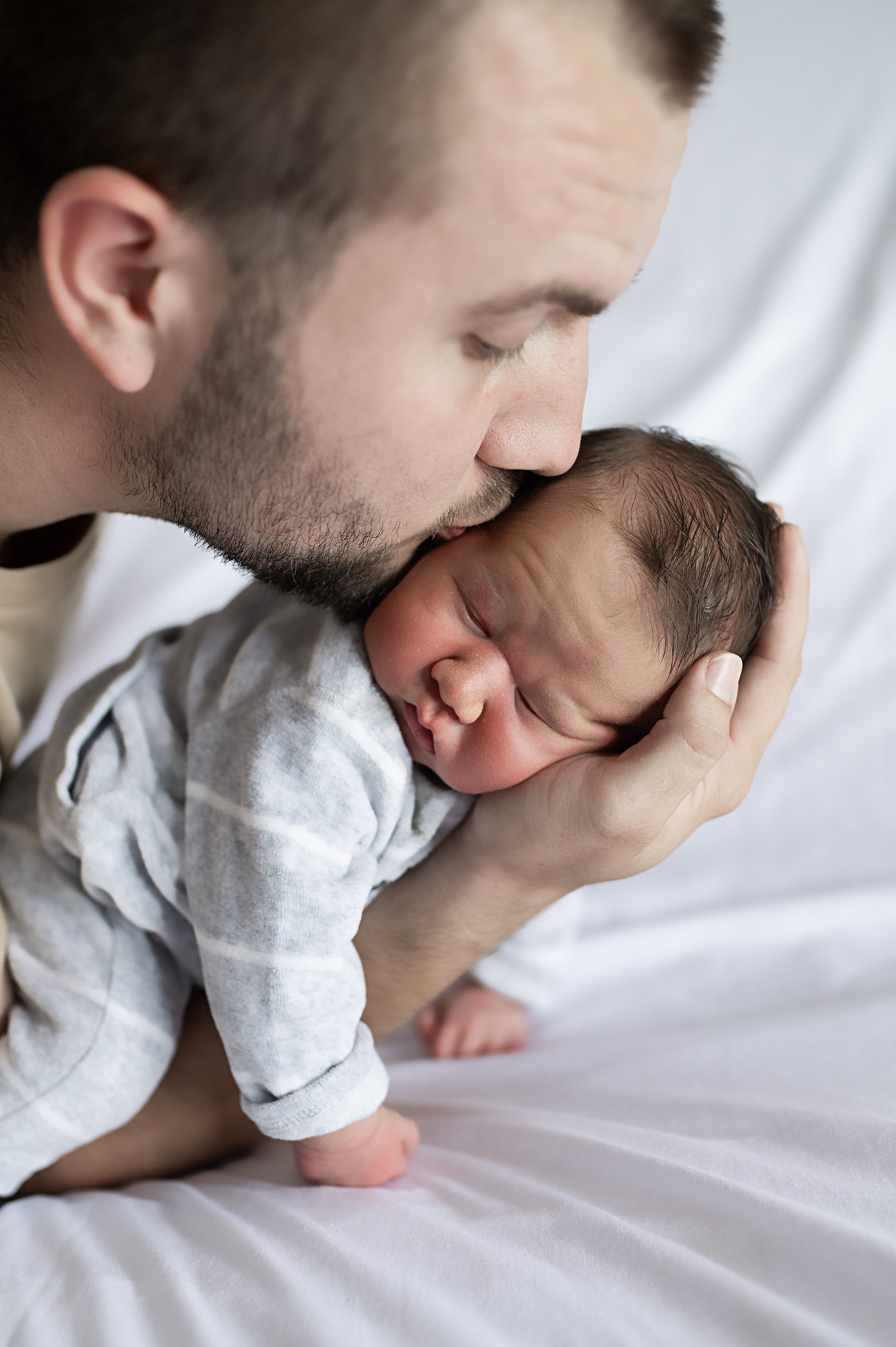 Un homme avec une barbe embrasse tendrement un bébé endormi, tenu dans sa main, sur un lit blanc.