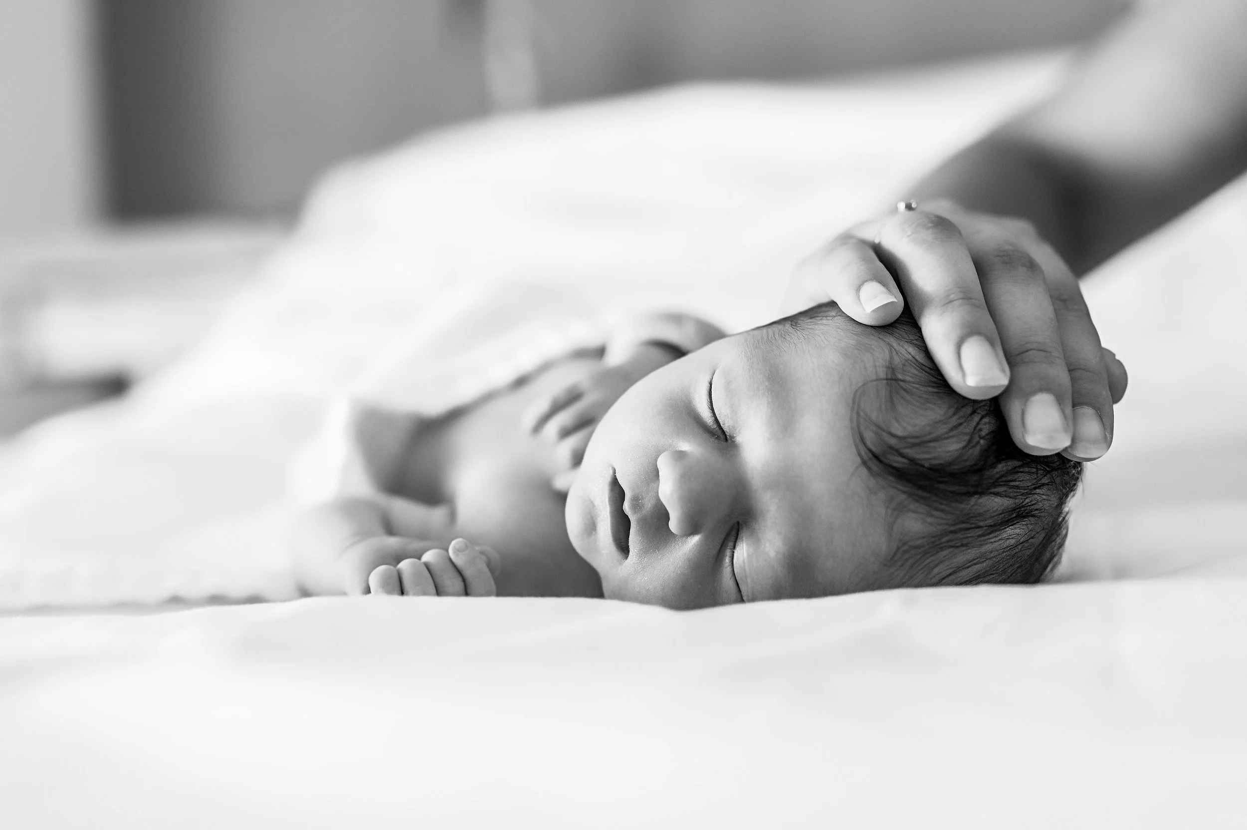 Un bébé dort paisiblement, une main humaine le caressant doucement sur la tête. En noir et blanc.