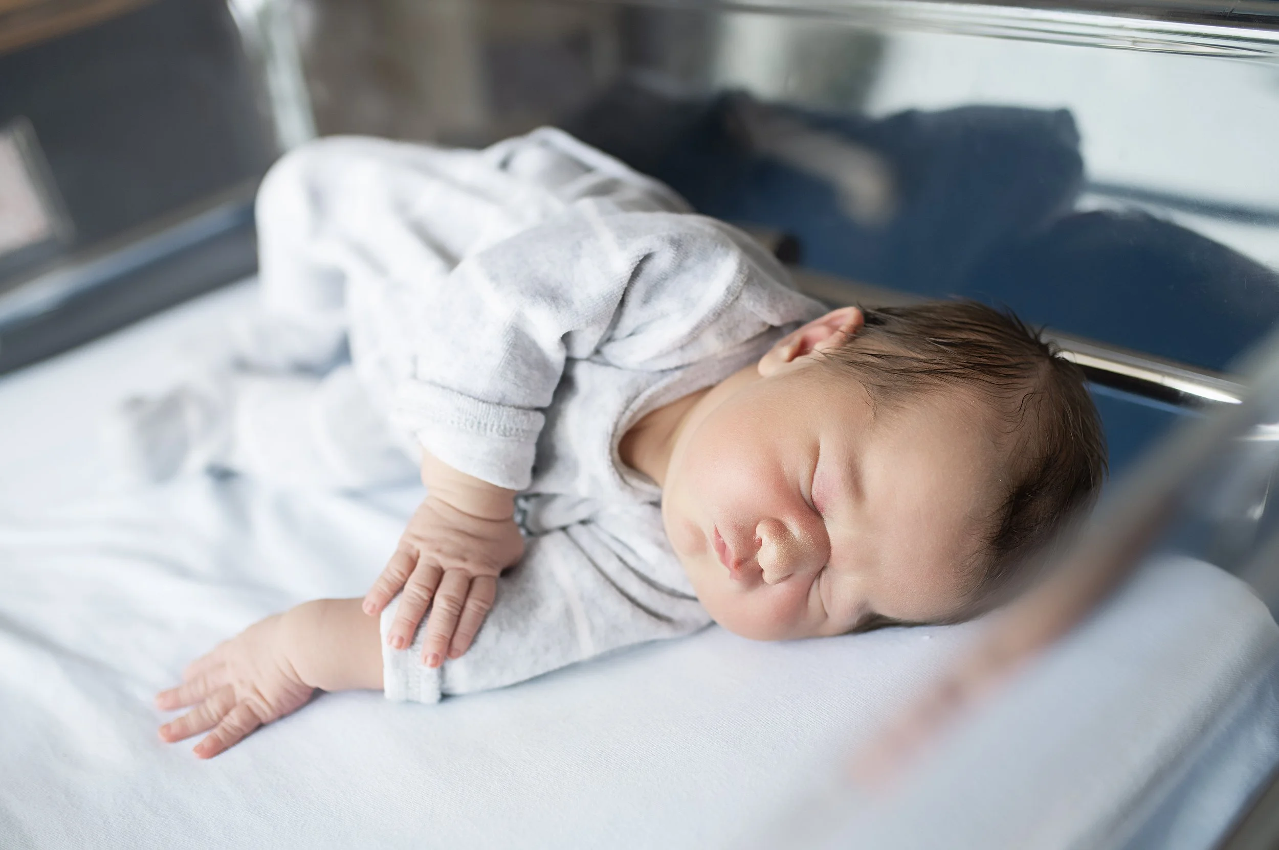 Un bébé dormant dans un lit d'hôpital avec un vêtement gris et une couche, allongé sur une couche blanche.